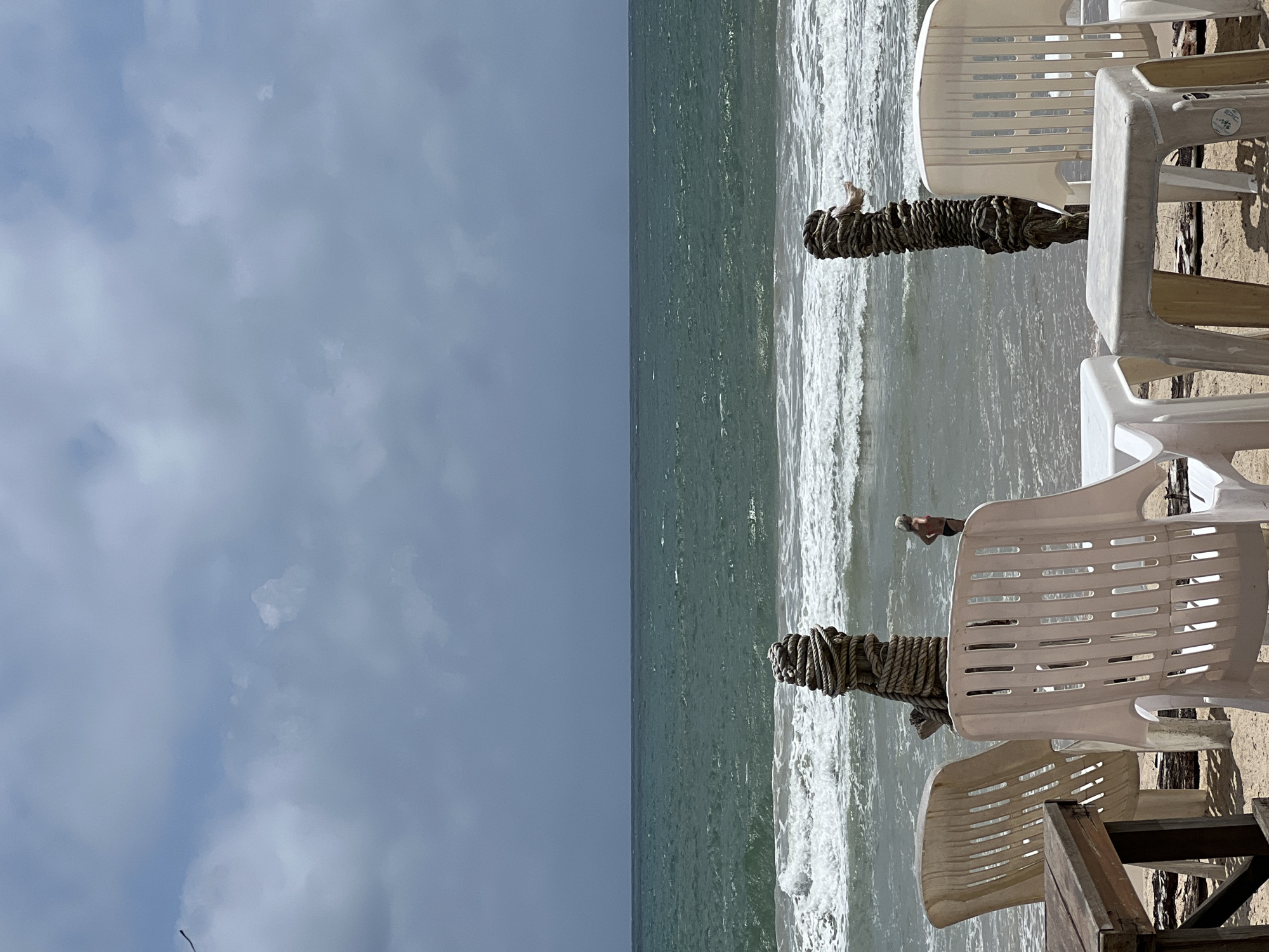The image depicts a beach scene in an unknown city. The left half of the image shows a cloudy sky. The right half presents the ocean with waves. The foreground contains numerous white plastic chairs. There are wooden posts wrapped in thick rope, standing near the chairs, and they mark the edge of the beach towards the ocean. Someone appears to be standing in the ocean. The composition suggests a beach resort or similar leisure area.