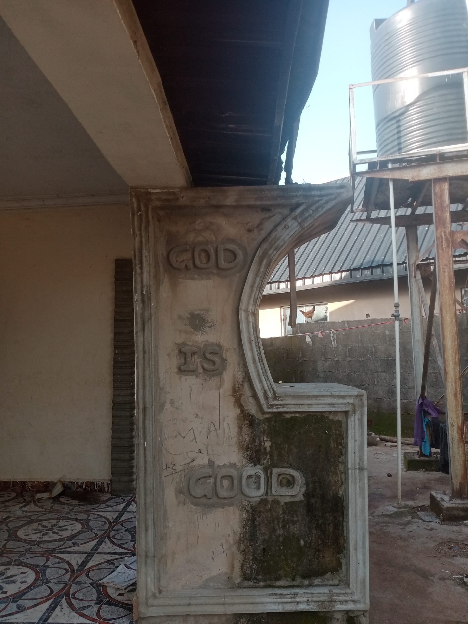 The image shows an exterior view of a concrete pillar with embossed lettering that reads "GOD IS GOOD". The pillar is weathered and stained, with some moss growth visible. To the left of the pillar is a wall with patterned tiles on the floor. In the background, a large water tank is visible on a metal platform, along with a corrugated metal roof and a clear blue sky. A chicken is perched on a wall in the middle ground. The scene appears to be in Jalingo, Nigeria, as indicated by the overall architectural style and environment, which is consistent with residential or commercial structures in the region.