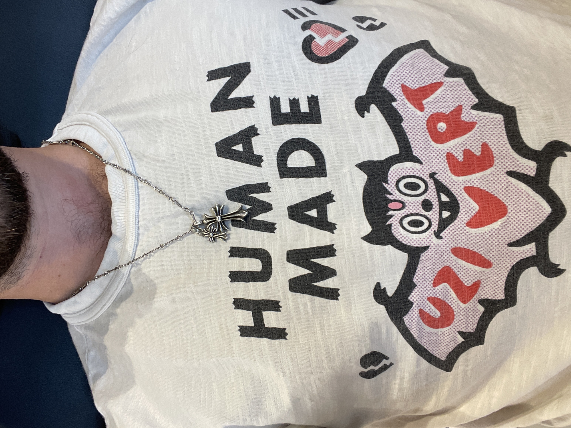 A person is lying on a blue surface, likely a couch, wearing a white t-shirt with "HUMAN MADE" printed vertically in black block letters on the left side of the shirt. A large cartoonish bat graphic is on the right side of the shirt, with a gray outline, pink wings, and red letters. A silver necklace with a cross pendant is visible around the person's neck. The person has a beard and appears to be of fair complexion. The image does not provide any information about the location being Mexico City, Mexico.