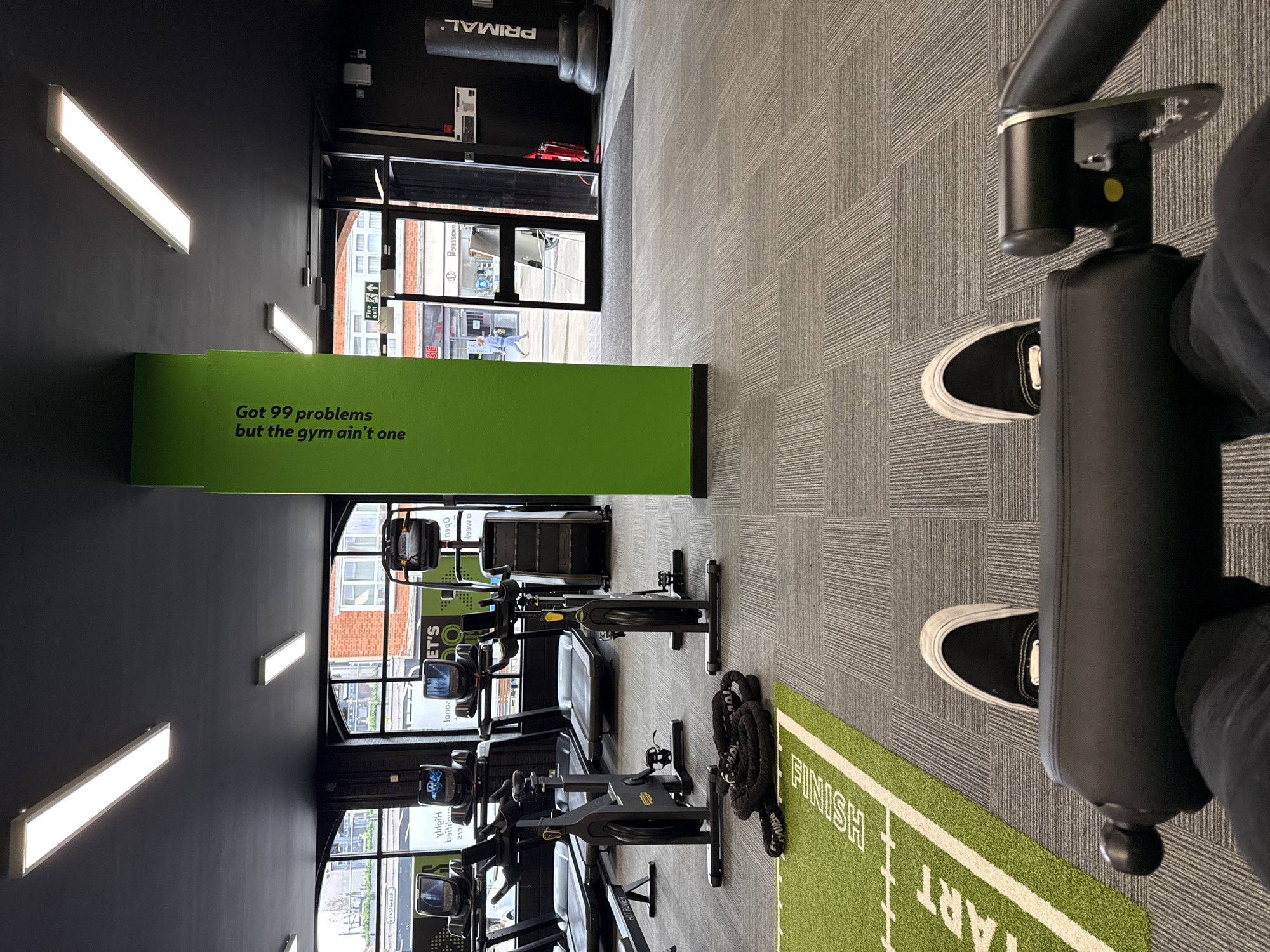 This is a high-angle, portrait-oriented photograph taken inside a gym in Letchworth, United Kingdom. A person wearing black and white sneakers and dark pants is seated on a piece of gym equipment. The camera's perspective gives the viewer a sense of being seated on the equipment. The gym is equipped with several elliptical machines and a treadmill, placed in rows along the wall. A green mat with the word "FINISH" and "START" printed in white is partially visible on the right side of the frame.
The gym has a modern design with a black wall. A green, rectangular display with the phrase "Got 99 problems but the gym ain't one" is visible along one wall. Long, rectangular lights are affixed to the ceiling.
A window to the outside world can be seen beyond the equipment. Buildings with brick facades, and a sign that reads "ECB PROFESSIONAL" are visible. The windows allow natural light into the gym. A "PRIMAL" branded heavy bag hangs from the ceiling in the upper left corner.