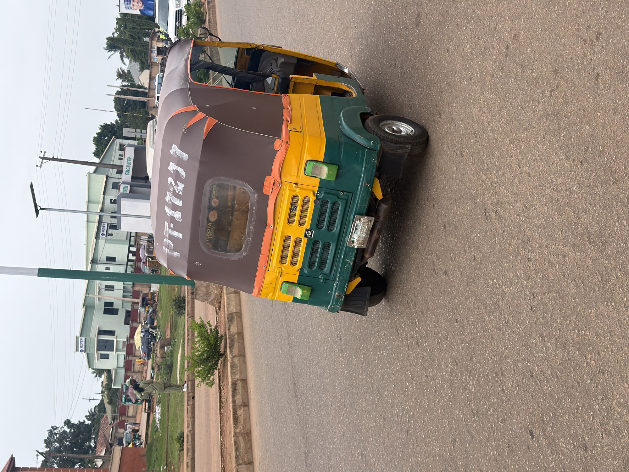 The media shows a side view of a colorful auto-rickshaw parked on an asphalt road. The auto-rickshaw is predominantly brown on top with a yellow and green section at the front. Orange trim is visible along the edge of the brown roof. The vehicle has a single visible wheel on the right side, and the rear section with its wheel is also visible. The license plate is partially obscured but appears to read "AKW 582 MH". The background reveals a street scene in Bukuru, Nigeria. Buildings, including one with a sign that says "MEDVIEW", are visible. There are streetlights and power lines indicating an urban or semi-urban setting. A few people are present in the background, including individuals in what appears to be military or security uniforms walking on a grassy area beside the road. The overall lighting suggests it is daytime, and the sky is overcast, indicating possibly cloudy weather. The scene appears to be a typical street view with a focus on the parked auto-rickshaw. Visible text includes "MEDVIEW" on a building and the license plate details on the auto-rickshaw.