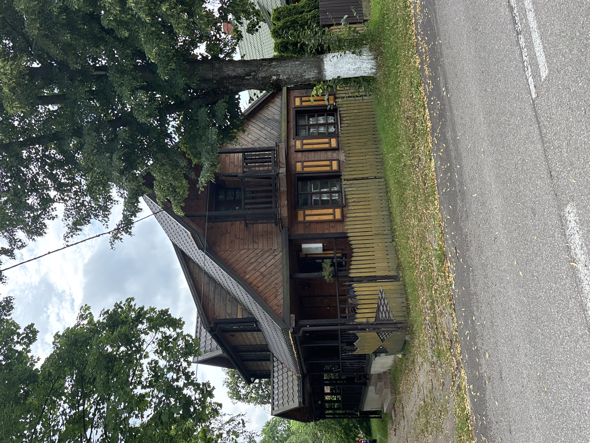 The image depicts a two-story wooden house with a dark gray roof, located in the village of Trześcianka, Poland. The house has a covered porch running along the front, supported by dark wooden posts. A light-colored wooden fence encloses the property in front of the house. The house features dark-trimmed windows, some with decorative shutters. A large tree stands to the right of the house, with its trunk painted white at the base. The foreground shows an asphalt road with some scattered leaves. The sky is overcast. No people are visible in the image.