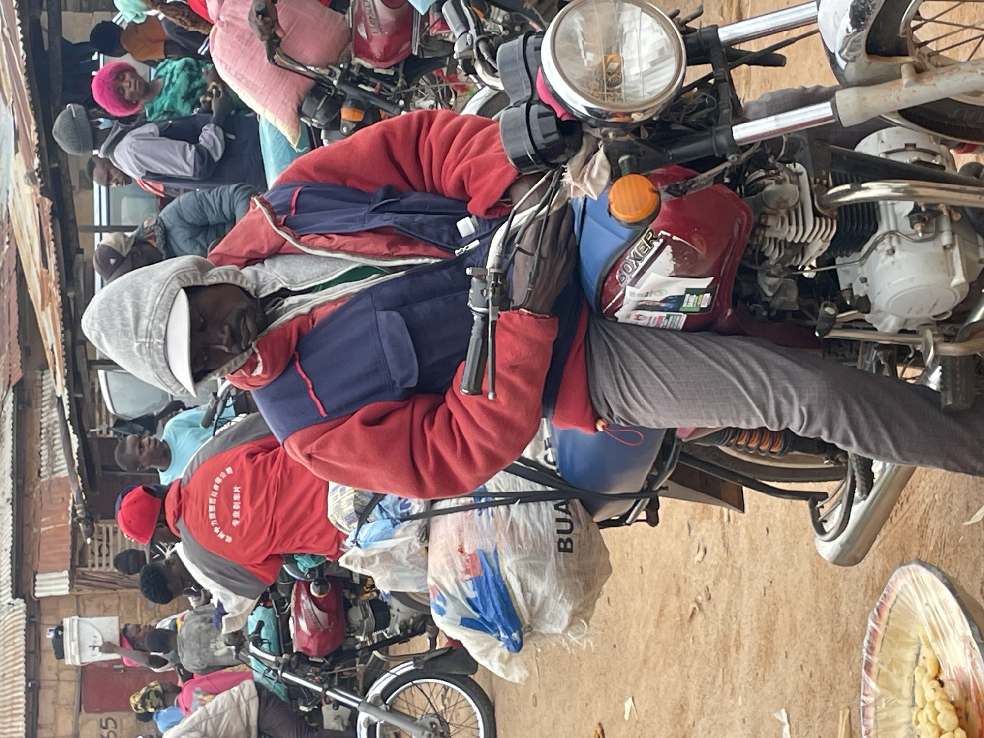 In Wang, Nigeria, a man wearing a red, blue, and gray hooded jacket, gray pants, and a gray baseball cap is seated on a red motorcycle. The motorcycle is equipped with a blue seat and a large white bag secured behind the seat with straps. The bag appears to contain other items, including a blue bag. The man is looking downwards and his hands are on the handlebars, suggesting he is either preparing to ride or has just stopped.
In the background, other people are visible, some standing and some seated on motorcycles, amidst what appears to be a bustling market or gathering. Structures with corrugated metal roofs are present, characteristic of informal settlements or market stalls. The ground is dusty and sandy. A bowl of yellow, twisty food items is in the foreground to the left of the motorcycle. The overall scene depicts a vibrant, everyday moment in a Nigerian community, likely within the city of Wang, given the context provided.