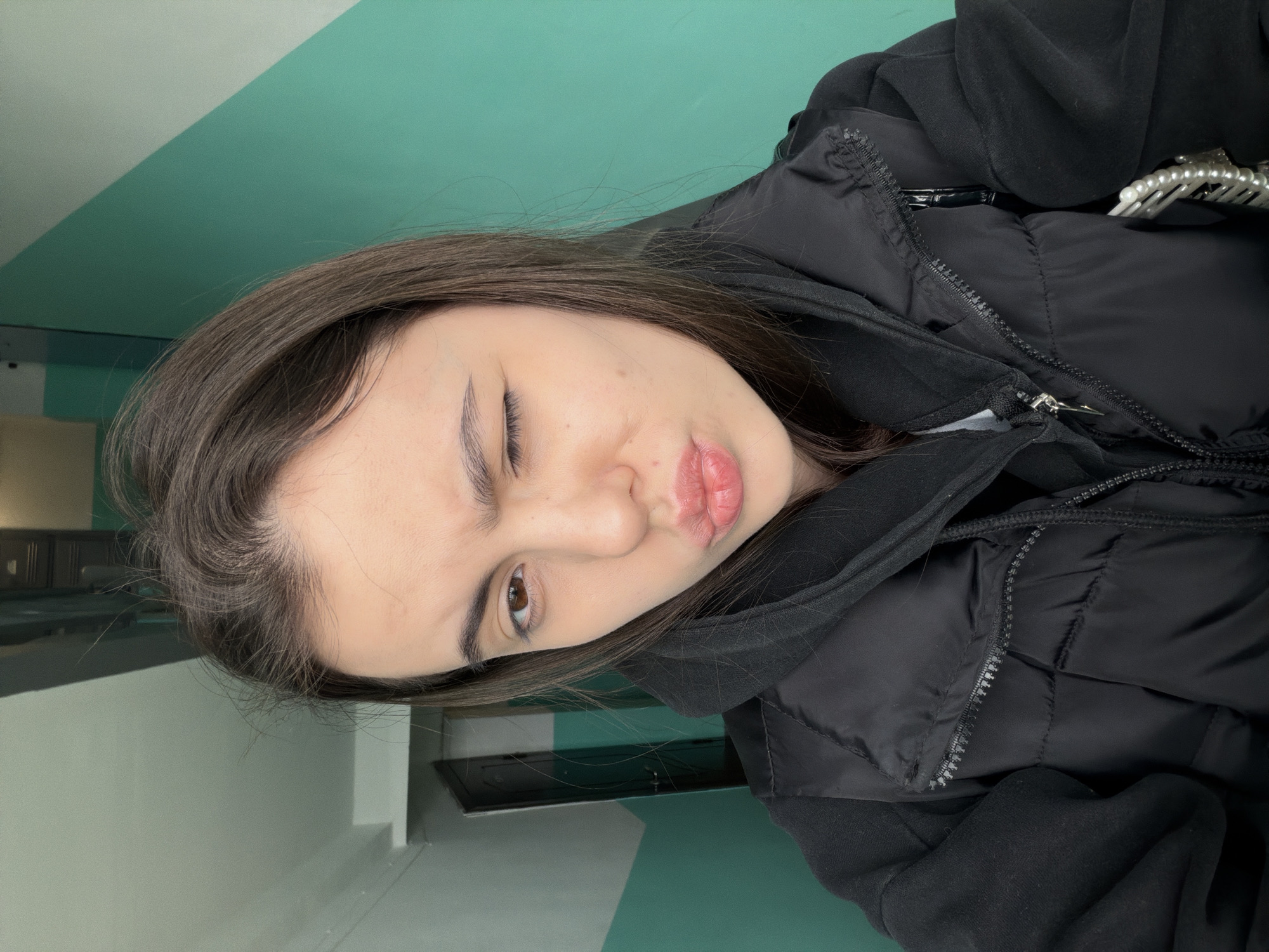 A young woman poses for a selfie in what appears to be an indoor location, possibly a public building due to the green and white striped wall. She is winking with her left eye and puckering her lips. She is wearing a black puffer jacket layered over a black hoodie. Part of her dark brown hair is visible. A hint of an object with pearls can be seen at the top right of the image near her jacket. The location is unknown, there is no visible information available to confirm it being in Chaikovsky, Russia.