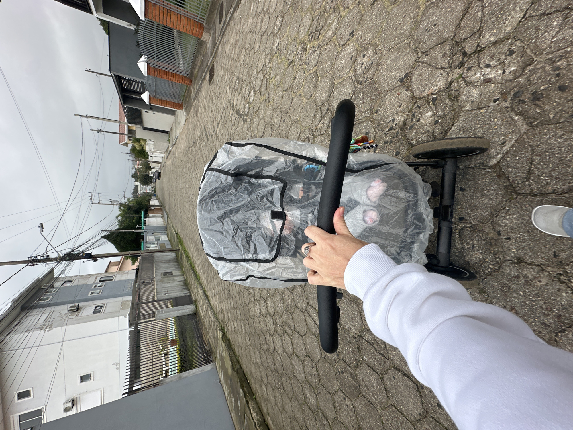 The image shows a first-person perspective of someone pushing a stroller on a cobblestone street lined with residential buildings and fences. The sky is overcast, suggesting cloudy weather. In the foreground, a hand wearing a white sweatshirt is gripping the stroller's handle. The stroller is covered with a clear plastic rain cover, and the feet of a baby, wearing pink socks, are visible inside. A car is parked further down the street. The architecture of the buildings suggests a suburban or urban residential area. The paving is made of interlocking hexagonal stones, common in some parts of Brazil. The scene depicts a casual walk with a baby in a stroller.
This image is likely from Florianópolis, Brazil, due to the distinctive hexagonal cobblestone paving, which is characteristic of many streets in the city, particularly in older neighborhoods. The architectural styles of the houses, a mix of modern and more traditional designs, are also consistent with residential areas in Florianópolis. The overall environment of a street with houses, fences, and utility poles is typical of a Brazilian city.