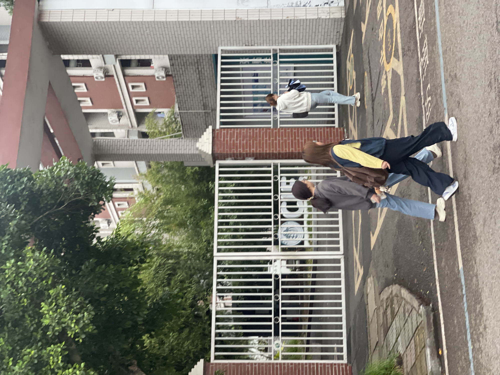 The image depicts a street scene outside a building in Chongqing High-tech Zone, China. Three individuals are walking on the pavement adjacent to a white metal gate and a brick structure. The first individual is wearing a white jacket and blue jeans. The second individual is wearing a yellow and blue jacket. The third person is wearing a gray jacket and blue jeans. Buildings are visible in the background. The pavement has yellow markings. Vegetation is also present in the foreground.