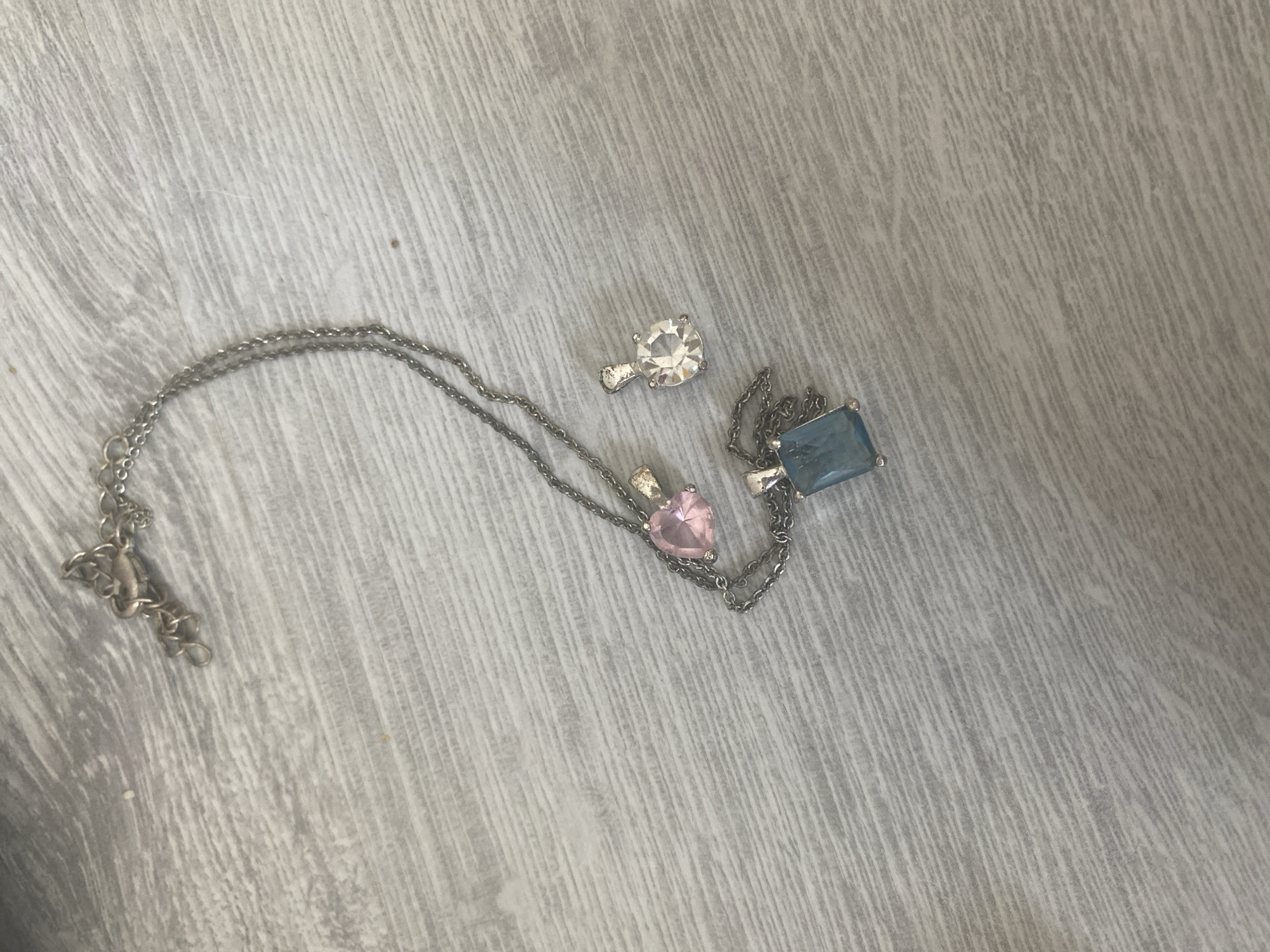 An overhead shot shows a silver necklace with three pendants on a light gray wooden surface. One pendant, a pink heart, is attached to the chain. Another pendant, a clear round stone, is separate from the chain. The third pendant, a rectangular blue stone, is also attached to the chain. The necklace is lying diagonally across the frame, with the clasp at the top left. The wooden surface has a prominent grain pattern. There are no people or any other objects visible in the image. The location of Blagoevgrad, Bulgaria is not evident from the image.