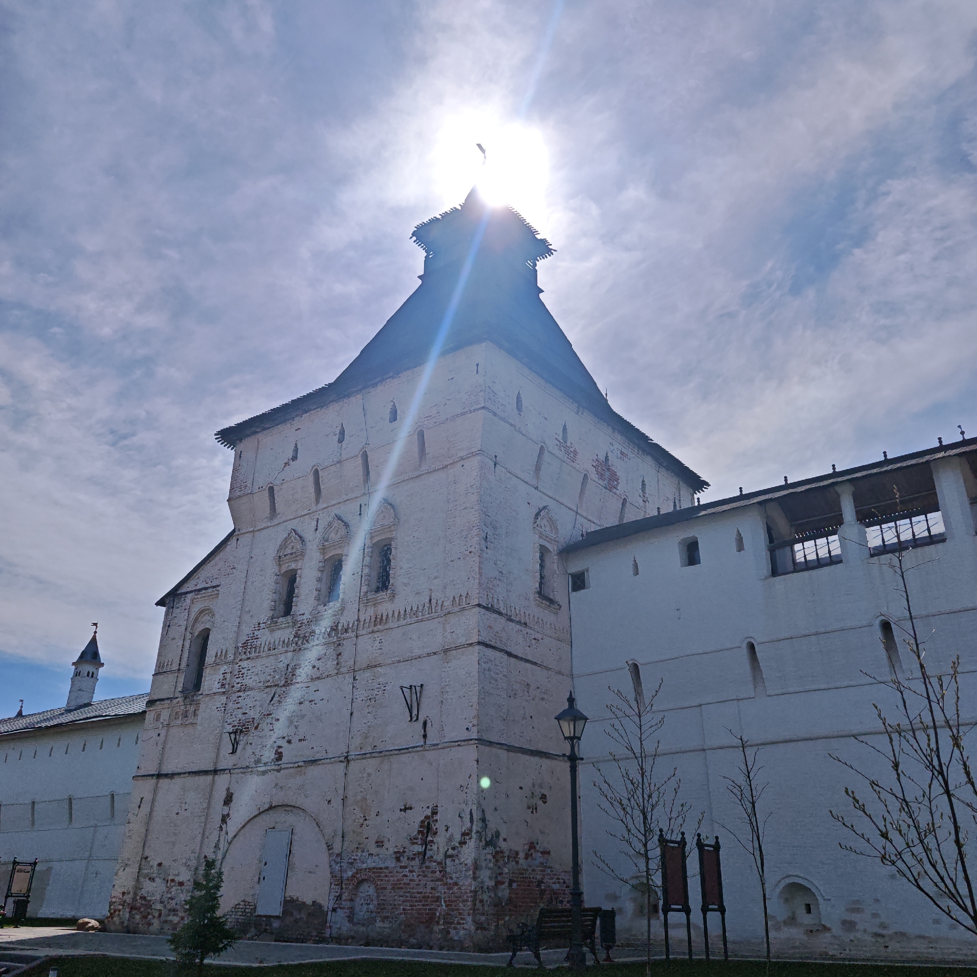 The image captures a tall, weathered white brick tower in Rostov, Russia. The tower, part of a larger architectural complex, features a black, sloped roof and a diminishing upper section. The sun is positioned directly behind the tower's peak, creating a lens flare effect with bright beams of light. The brickwork shows signs of age, with areas of exposed red brick. Adjacent to the tower is a long white wall with covered walkway. Bare-branched trees stand in front of the wall. In the foreground, there's a patch of green grass and a dark bench. A streetlight is visible near the base of the tower. In the distance, a pointed tower can be seen above the adjacent white wall. The sky is bright with a mix of clouds and blue patches.