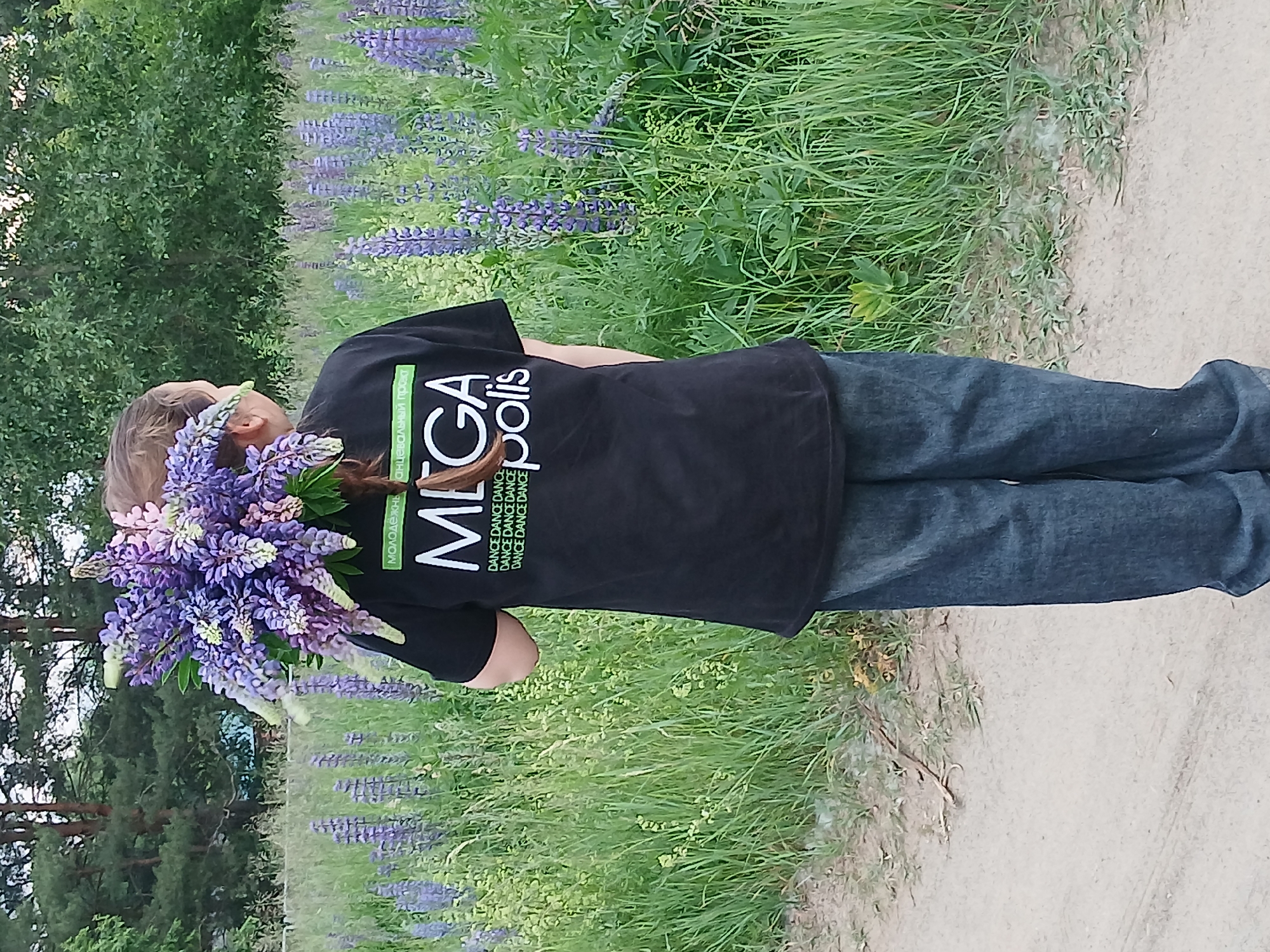 A person wearing a black t-shirt with white lettering and blue jeans stands in a field of lupine flowers, holding a bouquet of lupines over their head. The t-shirt has "MEGA" and "poliis" printed on it, along with smaller text that reads "МОЛОДЁЖНЫЙ ПРОЕКТ" and "DANCE DANCE DANCE" repeated. The person's hair is braided and woven with lupine flowers. The background consists of a dense green forest and more lupine flowers. The ground in the foreground is a sandy dirt path. The scene is consistent with a rural or park setting, possibly on the outskirts of the city of Chaikovsky, Russia, given the prevalence of lupine flowers often found in such areas during the warmer months.