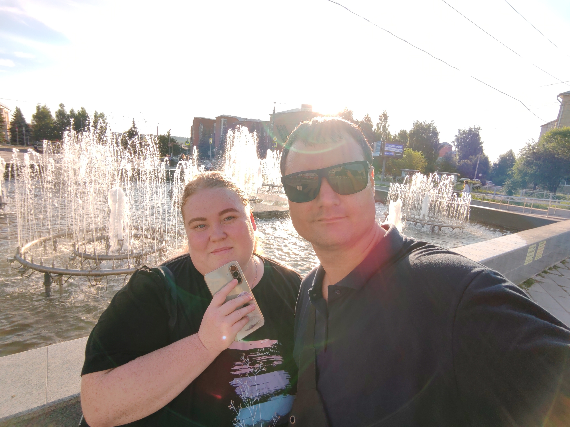 The media file is a selfie of a man and a woman in front of a large, illuminated fountain. The man, on the right, is wearing a dark polo shirt and sunglasses. The woman, on the left, is wearing a black t-shirt and holding a smartphone in her hand. She has a slight smile on her face. The fountain behind them is spraying water, creating a bright, sparkling effect in the sunlight. The setting appears to be an outdoor public space, possibly a park or a plaza, with trees and buildings visible in the background. The sun is low in the sky, suggesting it is either early morning or late afternoon. The overall mood of the image is cheerful and relaxed, with the couple enjoying a pleasant day out. There is no discernible text visible in the image.