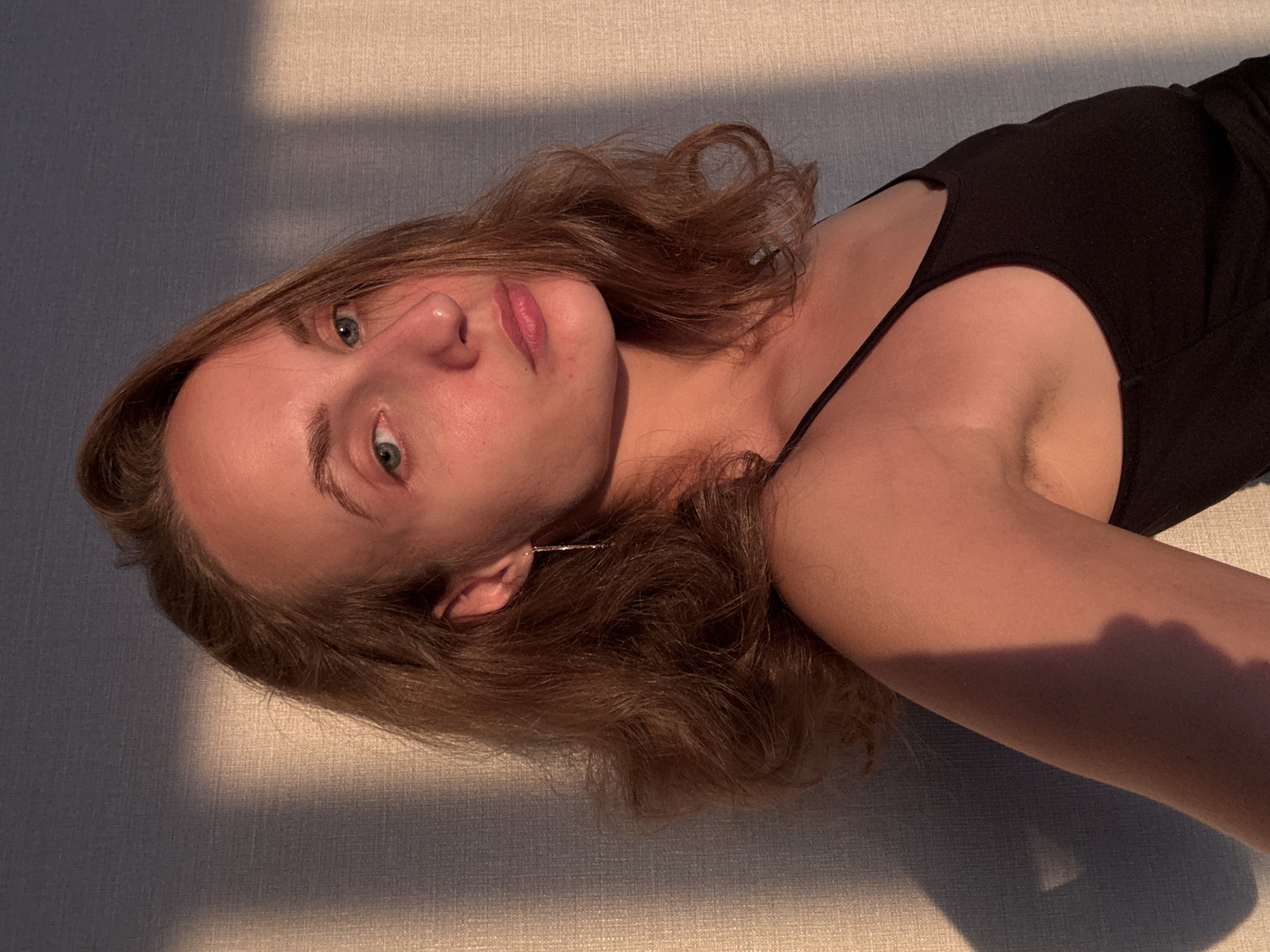 The media file shows a close-up, portrait-style shot of a young woman lying down, with her head tilted back and her body angled away from the camera. She has fair skin, light brown hair, and blue-green eyes. She is wearing a black tank top. The lighting suggests it is either early morning or late afternoon, with warm sunlight casting long shadows across her face and body, as well as the textured surface she is lying on. The background is a simple, textured fabric, possibly a couch or bed, with a plain wall behind it, showing a distinct shadow line. The overall mood is relaxed and serene. The image appears to be a selfie, given the perspective. There is no visible text. The location context is Podshivalovo, Russia.