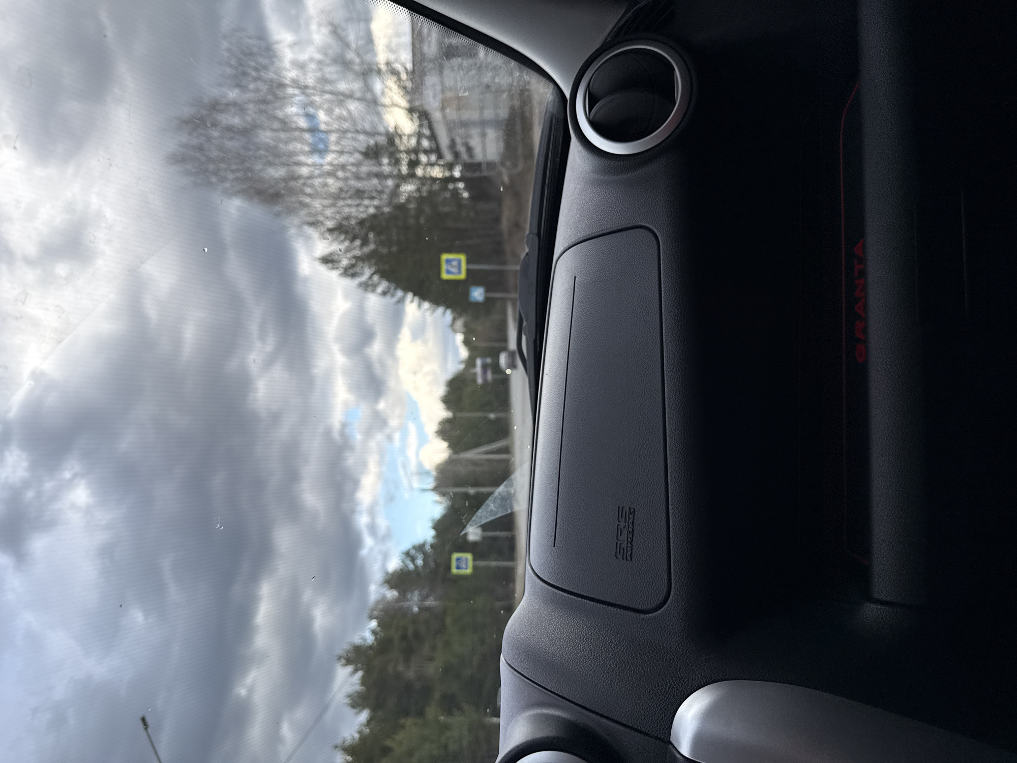 The image captures an interior view from a car, looking out through the front windshield onto a road scene. On the right, a portion of the car's dashboard is visible, featuring a circular air vent with a silver trim. Below the vent, a panel is marked with the text "SRS AIRBAG." Further to the right, a red, vertically oriented inscription reads "GRANTA," identifying the vehicle as a Lada Granta.
Through the windshield, an overcast sky with distinct cloud patterns and patches of brighter light is prominent. The windshield glass shows some water droplets or smudges. The external environment consists of a road flanked by trees, including both bare deciduous trees and some evergreens. In the mid-ground, two yellow square road signs with white pedestrian symbols are clearly visible, indicating pedestrian crossings. The presence of these signs and the general landscape suggest a road in a semi-rural or suburban setting. The time of day appears to be daytime, likely under an overcast sky or during the late afternoon. The overall scene depicts a mundane moment of travel or being parked on a road in Chaikovsky, Russia. No people are visible in the image.