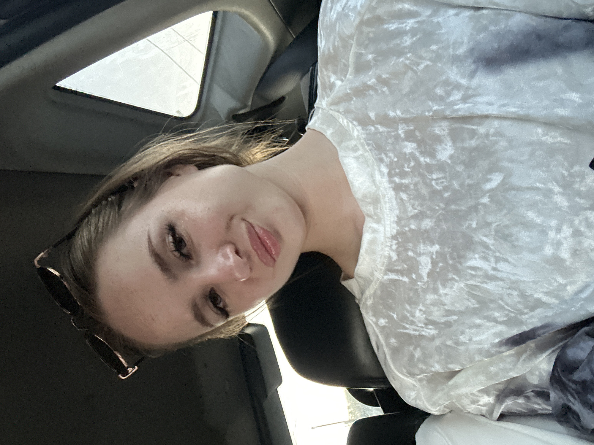 The image shows a young woman with blonde hair and makeup, wearing a light-colored, crushed velvet top. She is indoors, appearing to be in a car, with a sunroof visible above her. The woman is looking down and to the right, with a slight smile. Sunglasses are perched on her head. The lighting suggests it could be daytime. The overall mood is casual and relaxed. There is no visible text in the image.
