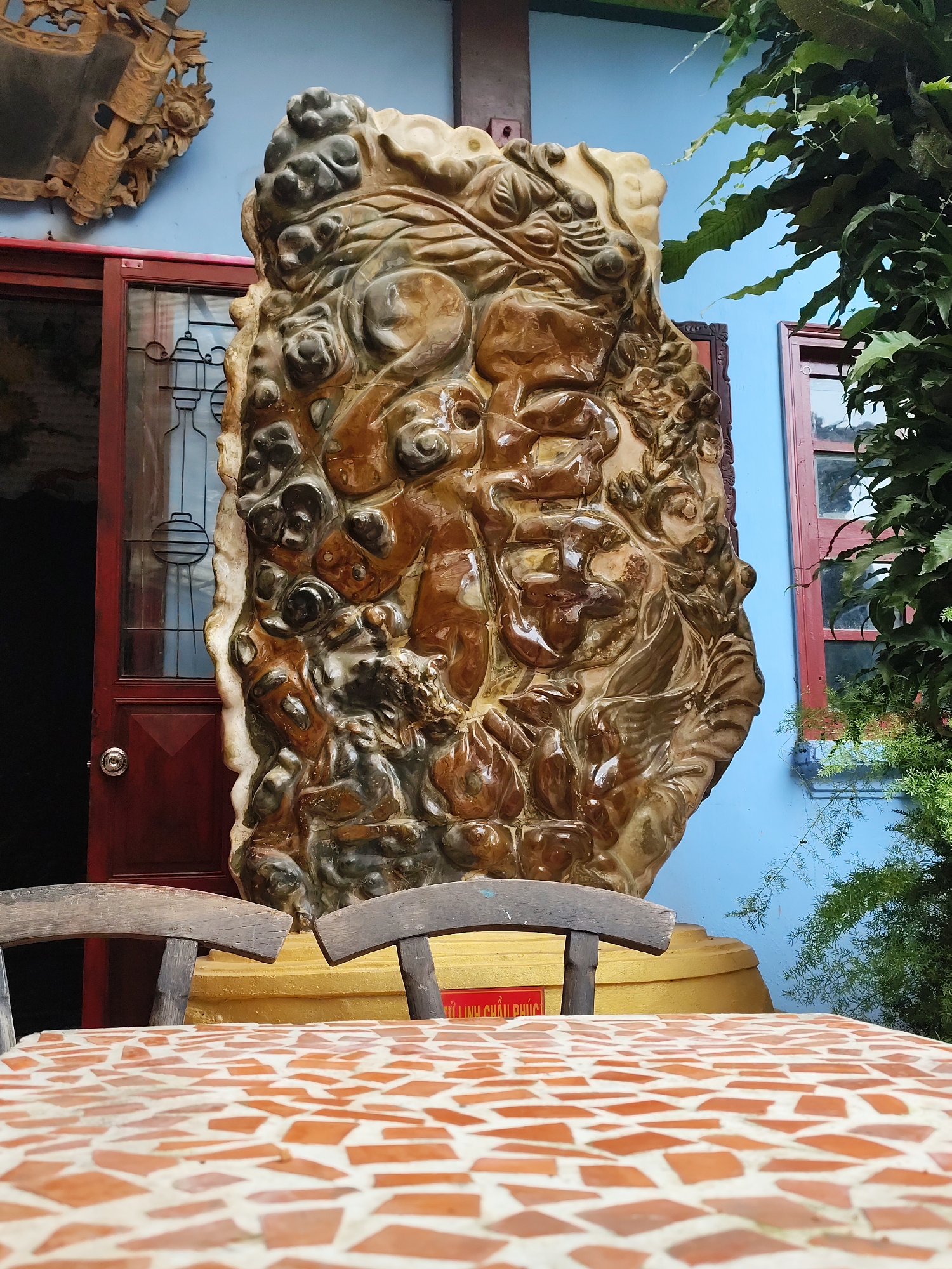 In Xuân Thành, Vietnam, a large, ornate, carved stone sculpture is displayed outdoors. The sculpture, primarily in shades of brown and beige, features a complex, irregular surface with what appear to be carved figures or natural formations. It stands on a golden pedestal. In front of the sculpture, a mosaic-tiled table with a terracotta and white pattern is visible, with two wooden chairs positioned behind it. The background consists of a bright blue wall, a red-framed window, and lush green foliage. To the left, a dark red door with glass panels and a circular silver doorknob is partially open. Above the door, a carved wooden decorative piece is affixed to the blue wall. The scene is set in what appears to be a courtyard or outdoor seating area.