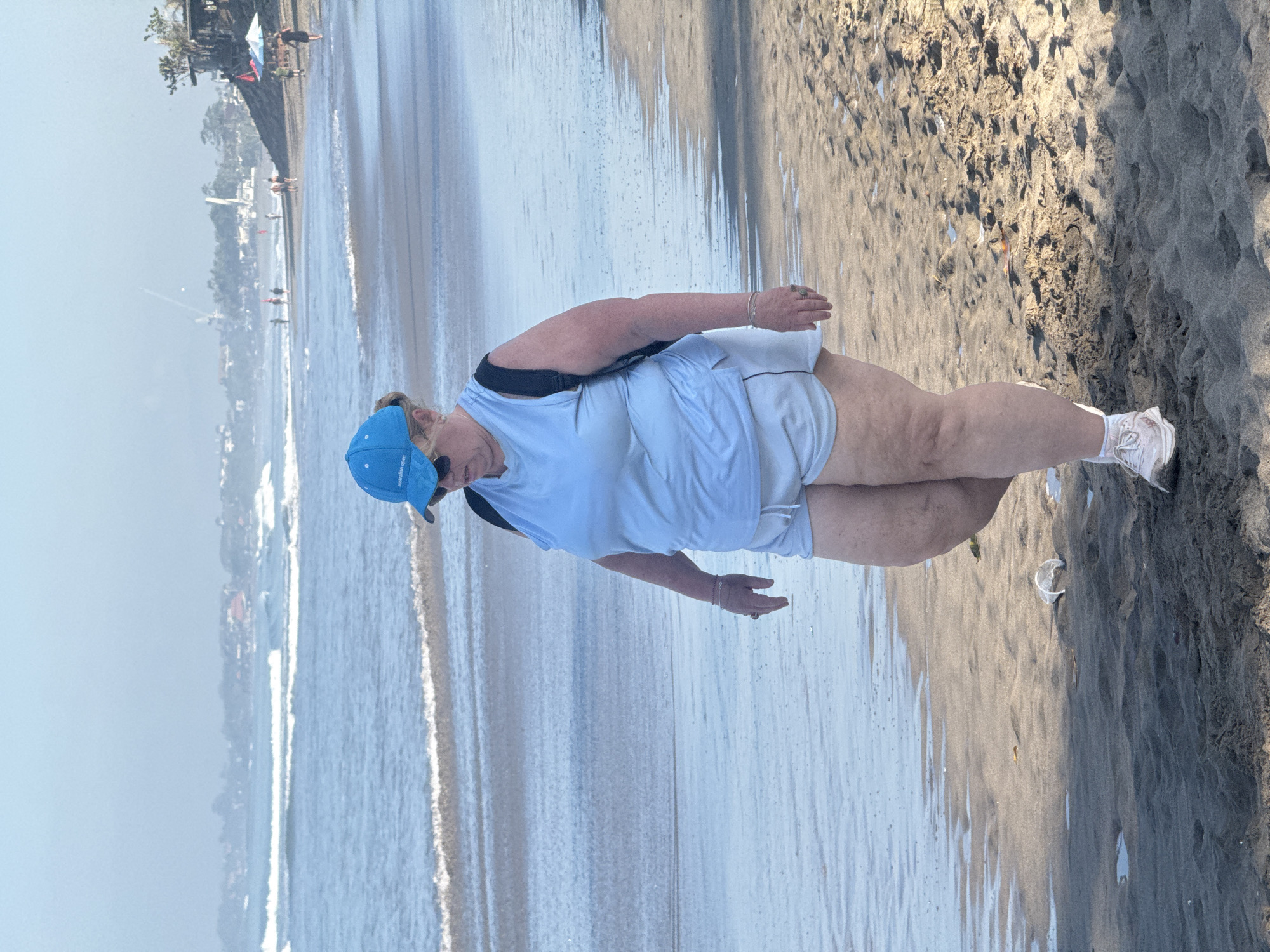 The image shows an adult woman standing on a beach in North Kuta, Indonesia. She is wearing a light blue cap, white shirt and shorts, and sneakers. She has a dark strap over her shoulder. The beach is sandy and wet from the receding tide. In the background, there is an out-of-focus view of buildings and people along the coastline.