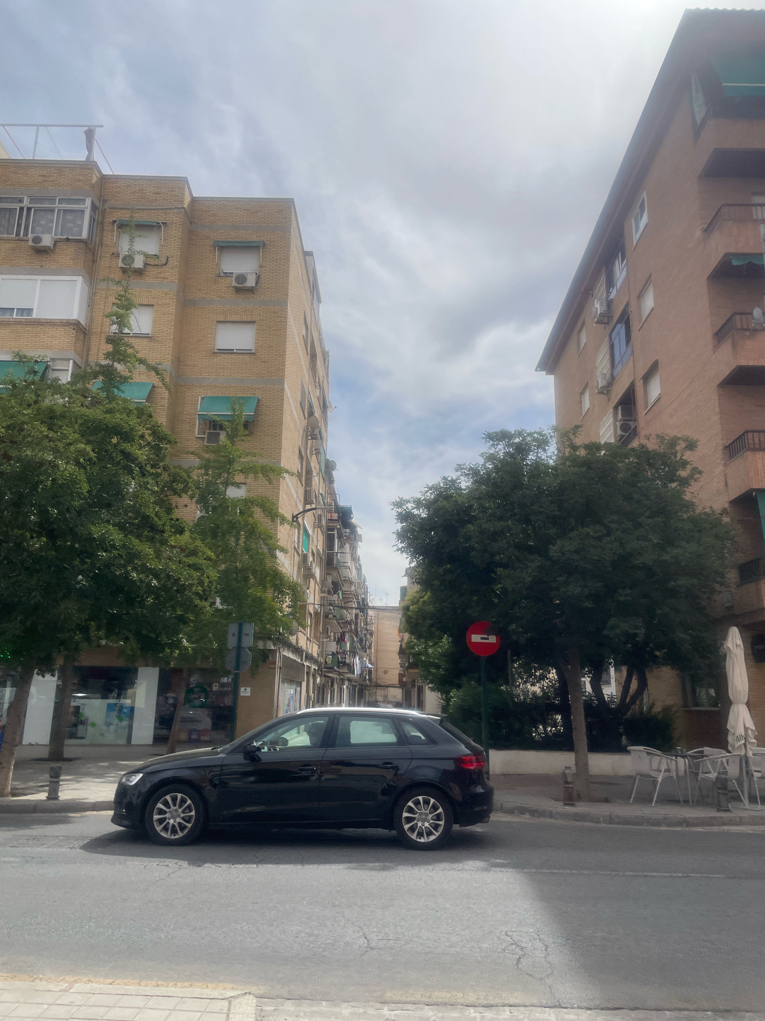 The image shows a street scene in Granada, Spain. A black Audi A3 hatchback is parked on the right side of the street, facing right. The car is in front of a light brown apartment building. To the left of the Audi, there is a light yellow brick apartment building with several air conditioning units visible on its facade. Two trees with green foliage are planted on the sidewalk in front of the yellow building. A red and white circular traffic sign is visible on a pole between the two apartment buildings. The street is paved and a sidewalk is visible on the left and right sides of the street. The sky is cloudy. Outdoor seating with tables and chairs is visible in front of the light brown building, along with a white umbrella.