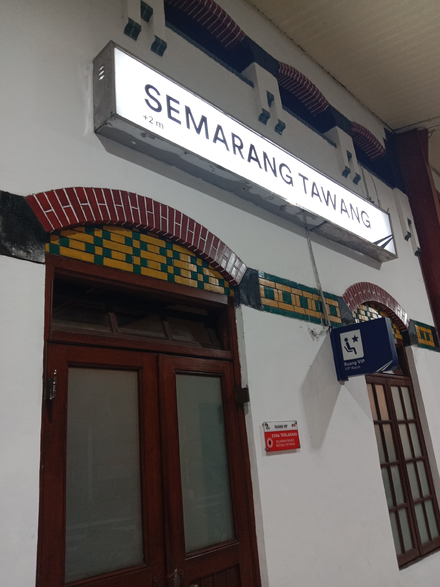 The image depicts the facade of a building in Semarang, Indonesia, likely a train station given the signage. A prominent illuminated sign displays "SEMARANG TAWANG" in large, dark lettering, with "+2 m" underneath. The architecture features decorative elements above a large wooden double door with glass panes. To the right of the door, a blue sign indicates "Ruang VIP VIP Room" with an icon of a person sitting and a star. Below the "Ruang VIP" sign, a smaller red and white sign reads "ZONA TERLARANG" and "DILARANG MASUK KECUALI PETUGAS," meaning "Restricted Zone" and "Entry Forbidden Except for Staff." The wall is painted white, with decorative brickwork arches above the windows and doors. The overall scene suggests an entrance to a public building with restricted access areas.