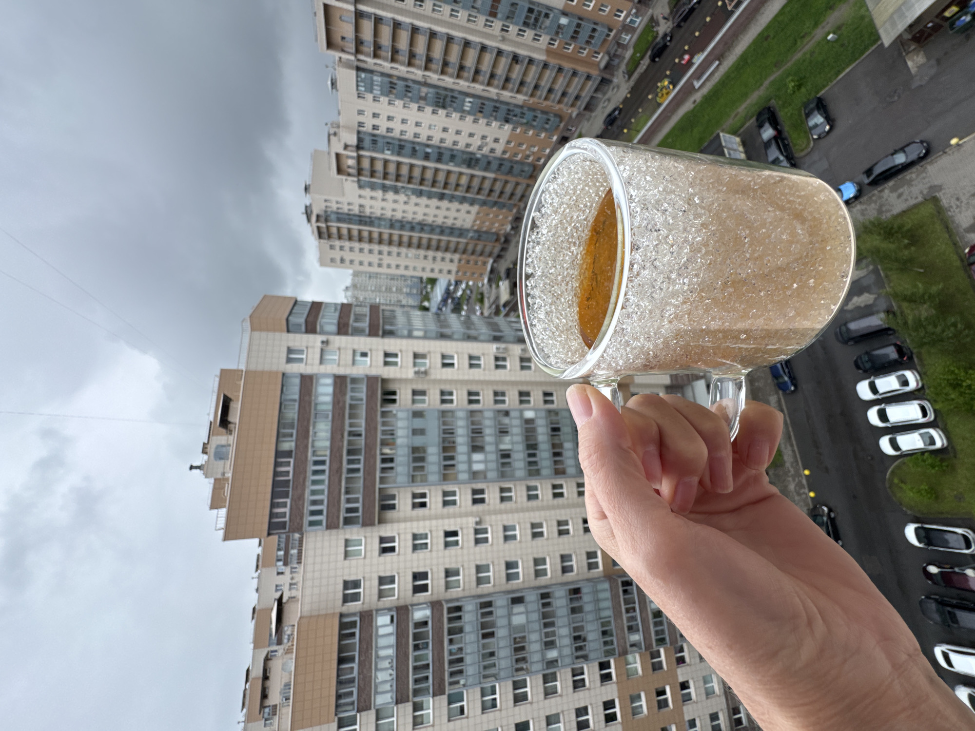 A hand holds a mug of tea against a backdrop of tall buildings, a street scene, and an overcast sky. The buildings are predominantly beige and gray, with rows of windows suggesting residential or office use. The street below is lined with parked cars and some patches of greenery. The sky is filled with heavy, gray clouds, indicating an overcast day. The mug, held in the foreground, is clear and appears to be filled with a light-colored beverage. The overall impression is that the photo was taken from a high vantage point, possibly a balcony or high-rise apartment, in an urban environment.