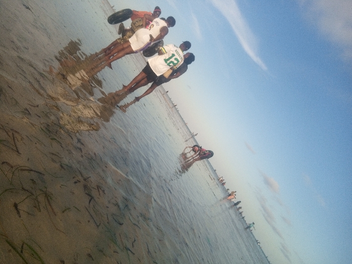 The image depicts a coastal scene in Shanzu ward, Kenya, featuring multiple individuals wading in shallow, clear water under a blue sky. In the foreground, three males stand partially submerged, with water reaching approximately their lower calves. The leftmost male wears an orange shirt and dark shorts, holding a dark, possibly deflated, tire inner tube. The central male wears a white basketball jersey with "LAKERS" and the number "3". The rightmost male wears a white and green basketball jersey with "RODGERS" and the number "12". Their reflections are visible on the water's surface, which contains sparse submerged seagrass. Further in the midground, another individual is seen partially submerged, also holding a dark tire inner tube. In the distant background, a horizontal line of numerous people extends across the water, indicating a populated shallow beach or tidal flat environment. The sky is predominantly clear blue with a few thin white clouds. The lighting suggests daytime.