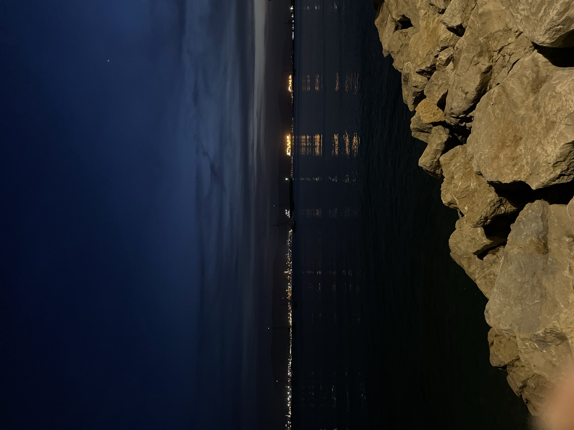 The image depicts a nighttime view of the sea from a rocky shoreline in La Línea de la Concepción, Spain. The sky is dark blue with faint clouds and a single bright star visible. Across the water, the distant city skyline is illuminated with numerous lights, creating reflections on the calm sea. Several large ships, also lit, are present on the water. In the foreground, a collection of large, textured rocks forms the edge of the land. The overall scene is peaceful and atmospheric, characteristic of a coastal city at night.