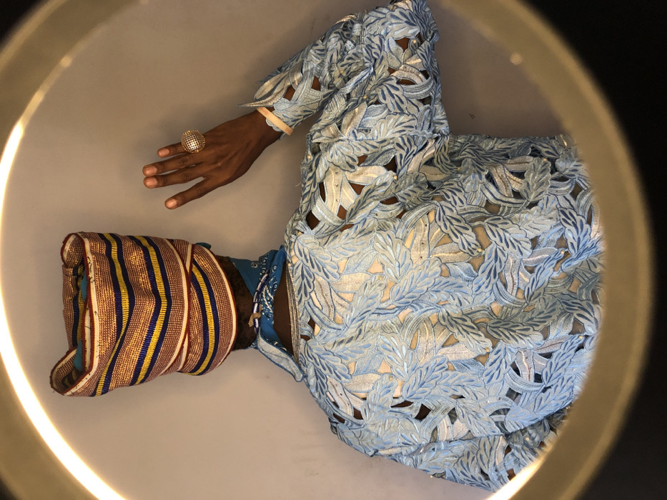 The image, captured in Ngleshi Amanfro, Ghana, displays the posterior view of an individual positioned against a plain, light-colored wall, partially encircled by the bright arc of a ring light. The person wears a structured headwrap composed of tightly folded fabric with alternating horizontal stripes in gold, blue, and red. A blue bandana is visible around the neck, beneath which a blue beaded necklace rests. The upper torso and right arm are clad in a light blue garment featuring an intricate pattern of cut-out leaf shapes, some edged with lighter blue or silver embroidery and adorned with small, reflective embellishments. Through these cut-outs, a warm-toned underlayer or skin is visible. The individual's right hand is raised, palm facing outward, with fingers slightly parted. A large, circular, gold-toned ring with a textured, mesh-like surface is prominent on the ring finger of the right hand. A pale, flat wristband is also present on the right wrist.