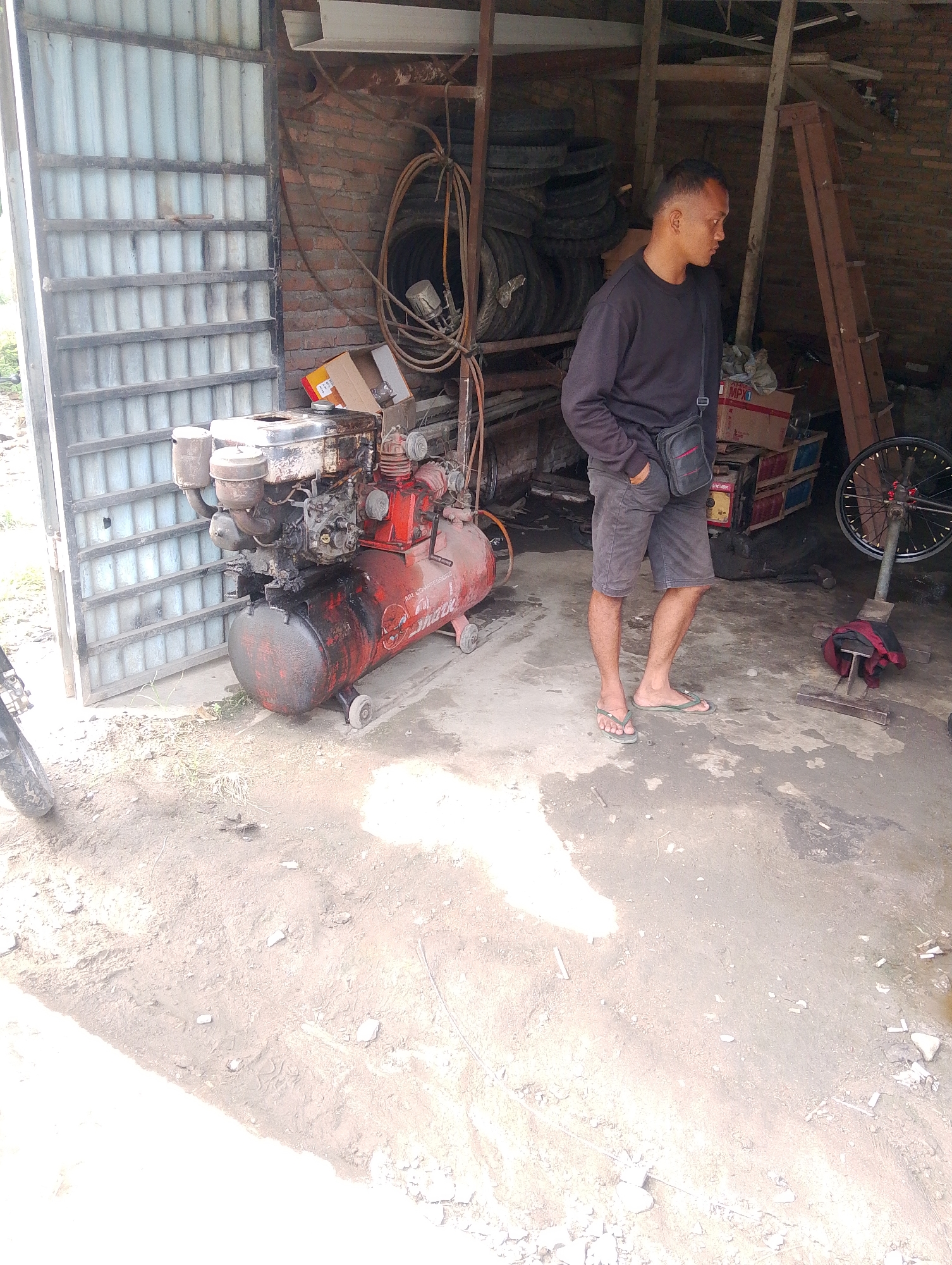 The image shows a man standing in what appears to be a workshop or garage. The setting is rustic, with a corrugated metal door on the left, brick walls in the background, and a dirt floor.

In the foreground, a large red air compressor with an engine on top is visible. The compressor is old and appears well-used, with some oil stains and dirt. A bicycle wheel is mounted on a stand to the right of the man. There are also stacks of tires in the background and various tools and equipment scattered around.

The man is wearing a dark long-sleeved shirt, dark shorts, and flip-flops. He has a small bag slung across his chest. He is looking down and to his right, with one hand in his pocket.

The overall impression is of a working environment, possibly a tire repair shop or a mechanic's garage, located in Naga Jaya 1, Indonesia. The lighting suggests it is daytime, with sunlight casting shadows.