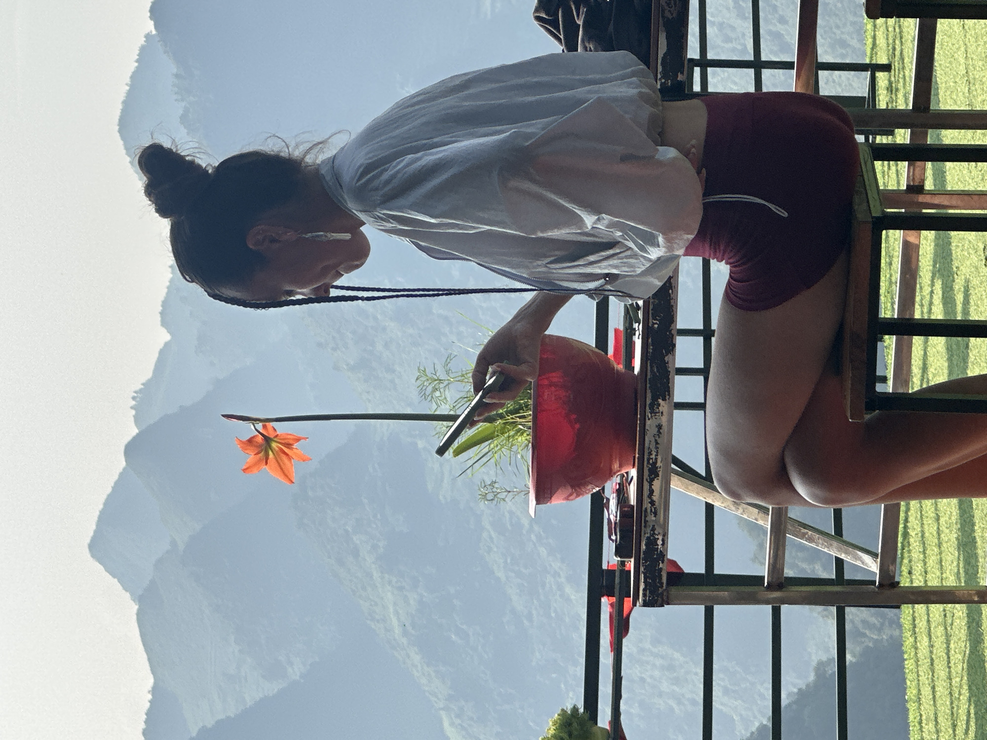 A woman with her hair in a bun, wearing a white top and red shorts, is kneeling next to a red pot containing plants, possibly on a balcony or raised platform in Đường Thượng Commune, Vietnam. She appears to be tending to the plants, holding a dark object, perhaps a tool or device, near the foliage. The background shows a mountainous landscape under a bright sky. The location can be identified because it is the only place that is called Đường Thượng Commune. A single orange flower blooms nearby. A black metal railing is visible in the foreground.