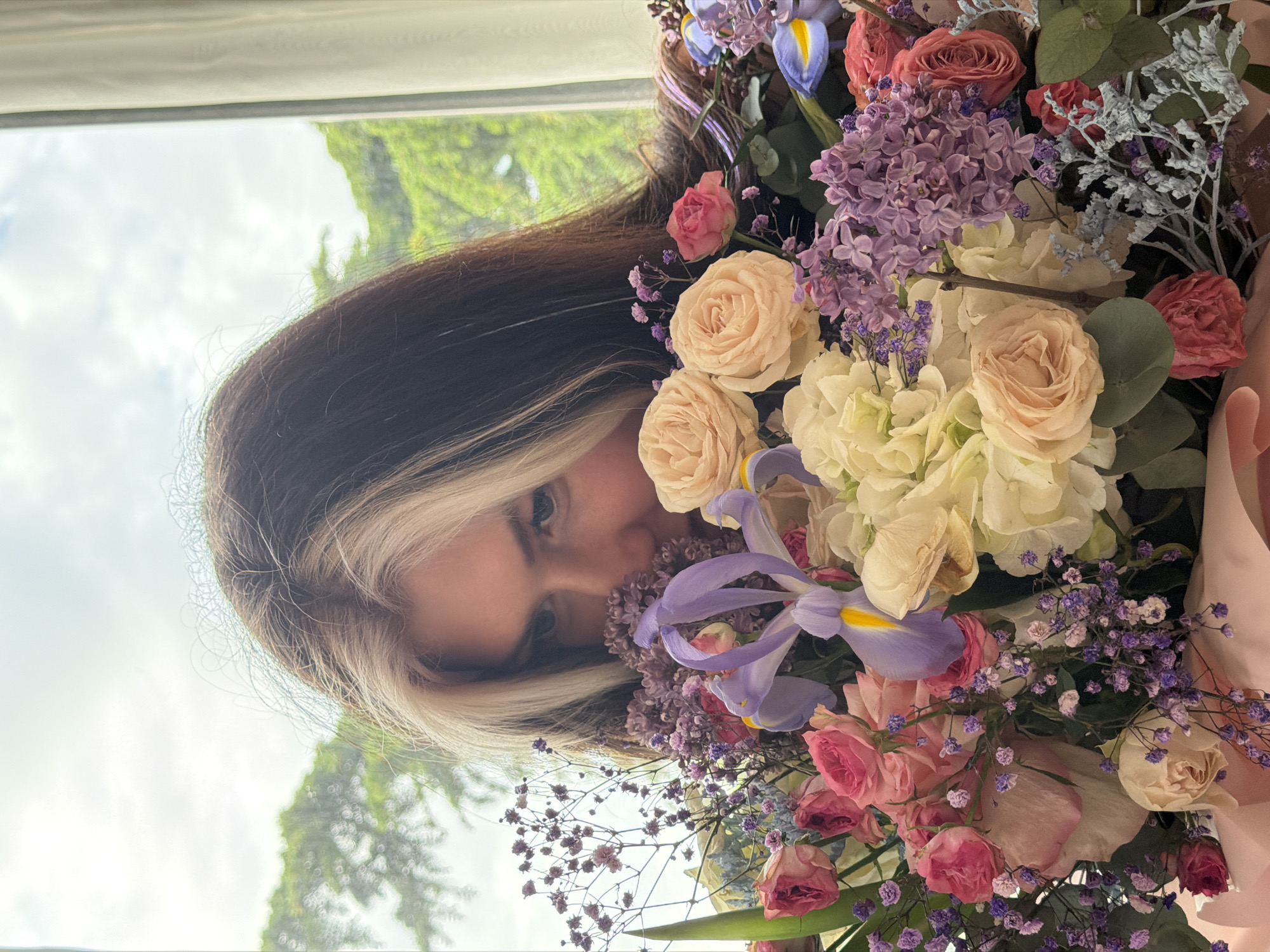 A sideways medium shot captures a person partially obscured by a large, vibrant floral bouquet, positioned indoors near a window in Budva, Montenegro. The person, likely female, has dark brown hair with distinctive blonde or white streaks framing her face, and her eyes are directed towards the viewer with a gentle, perhaps slightly smiling, expression as she peeks from behind the flowers.
The lavish bouquet dominates the foreground and features a rich assortment of blooms, including numerous light pink and peach garden roses, dense clusters of purple lilac, large white or cream hydrangeas, and striking purple irises with yellow centers. Smaller purple filler flowers are also interspersed throughout the arrangement. The bouquet is wrapped in a soft, light pink material or paper on the right side.
Through the window in the background, a bright, cloudy sky is visible, along with blurred green foliage from trees or bushes, indicating that the photo was taken during daylight hours. The overall scene conveys a cheerful and appreciative mood, focusing on the beauty of the flowers and the individual presenting or receiving them. There is no visible text in the image.