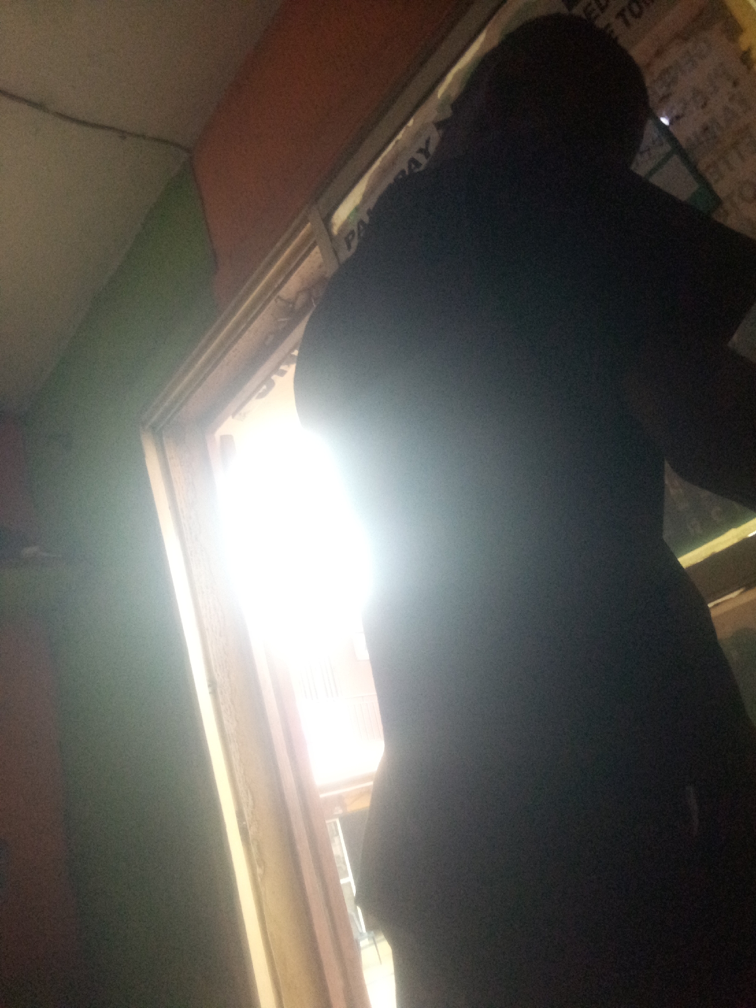 A full-body shot from a low angle captures a person silhouetted against a bright light source, possibly a doorway or window, in Shangisha, Nigeria. The person, wearing dark clothing, stands in profile to the right. The background is dominated by the intense light, with some indistinct shapes visible beyond it. To the left of the figure, a wall painted in shades of green and orange suggests an interior space. The overall lighting is high contrast, with deep shadows and blown-out highlights, making precise details difficult to discern.