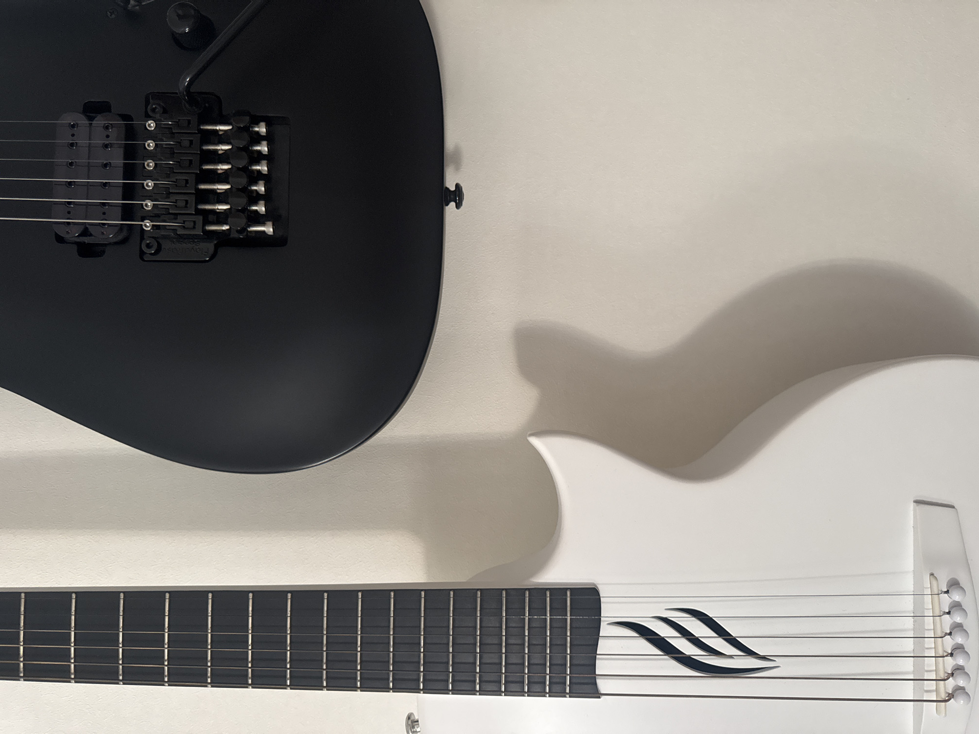 The image shows two guitars, one white and one black, hanging on a wall. The white guitar is in the foreground on the left side of the image, with its fretboard and body visible. The black guitar is in the background on the right, with its bridge and strings in focus. The guitars are positioned at an angle, with the white guitar partially obscuring the black one. The wall appears to be a neutral, light-colored surface, and a shadow is cast by the white guitar onto the wall, indicating a light source from the left. No people are present in the image, and no specific details about the location in Kremenchuk, Ukraine, can be deduced from the visual information provided.