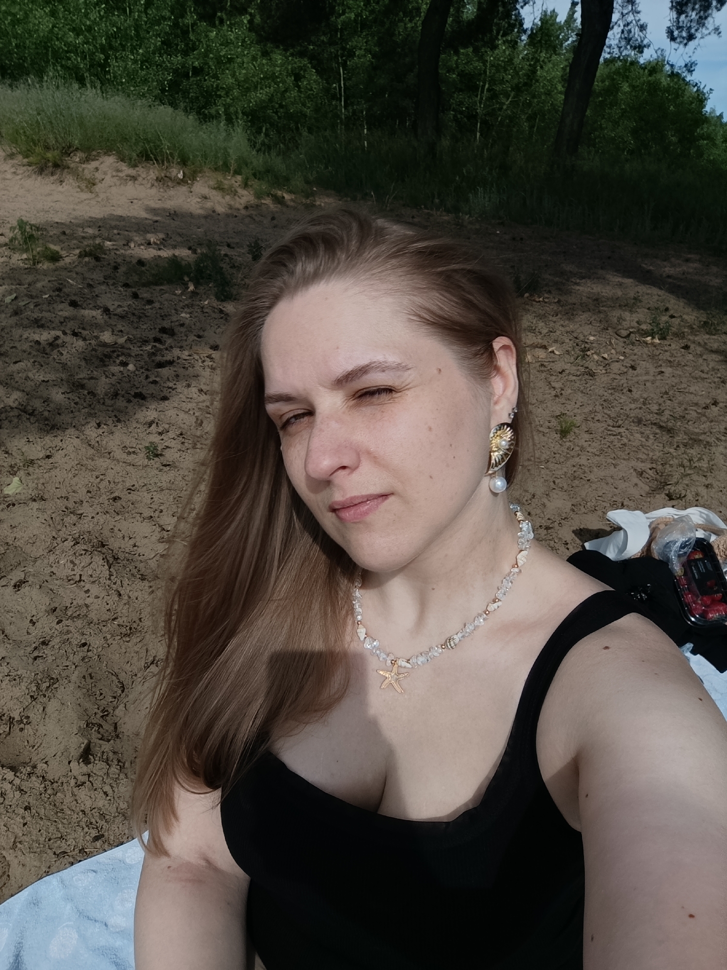 An outdoor, eye-level, medium shot shows a fair-skinned woman sitting on a sandy beach with trees and foliage in the background. The woman has light brown hair and is looking slightly off-camera to her left. She is wearing a black tank top, a shell necklace with a starfish pendant, and an earring shaped like a nautilus shell with a pearl. To her right, there is a black bag with white fabric and a clear plastic container filled with red berries. The sandy ground is visible in the foreground, with some dry leaves and sparse green vegetation. A light blue towel is partially visible beneath the woman on the left side of the frame. The lighting suggests it is daytime, with shadows cast on the ground.