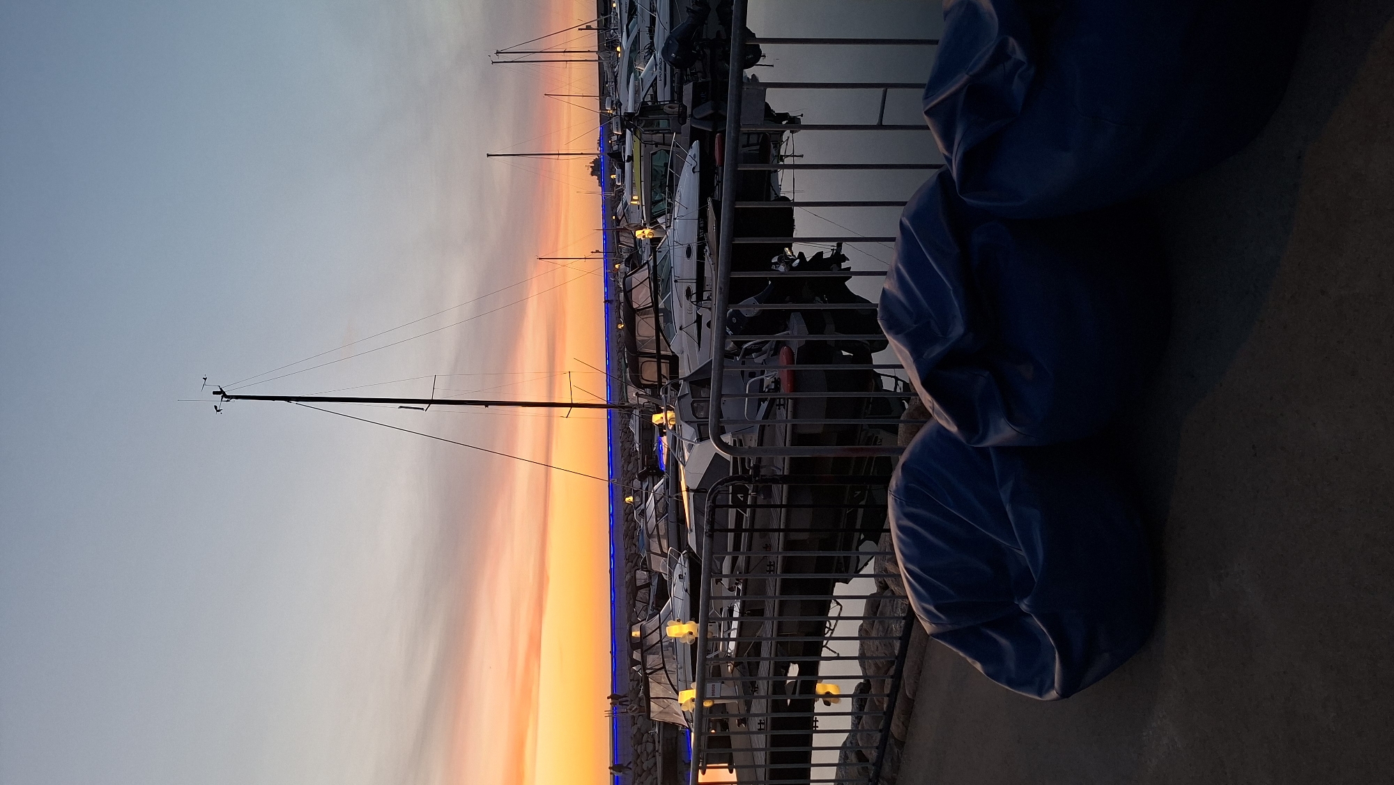 At dusk, a marina scene unfolds with a gradient sky of pale blue and orange hues. Several boats are docked, their silhouettes softened by the low light. A tall mast from a sailboat stands prominent against the sky, with wind measurement instruments at its apex. In the foreground, three dark blue beanbag chairs are arranged on a concrete surface, partially obscuring the view of the water and the docked vessels. The water reflects the muted colors of the sky and the lights from the boats. Along the shoreline, a line of blue lights is visible, illuminating a stone barrier.