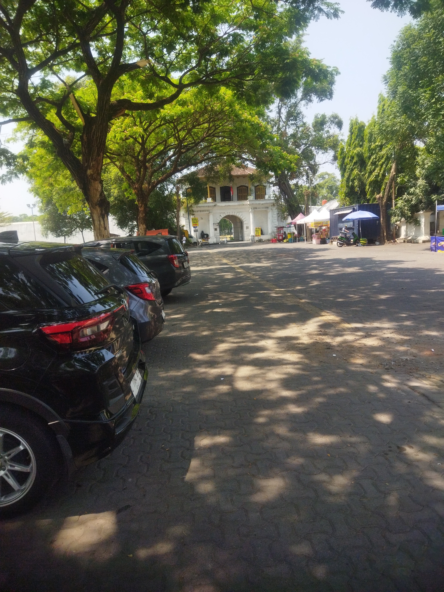 The image depicts an outdoor scene in Surakarta, Indonesia, featuring a row of parked black SUVs in the foreground. Beyond the vehicles, a white arched gateway, part of a historic building, is visible. Several temporary market stalls with tents are set up in front of the gateway. In the background, lush green trees dominate the scene, casting dappled shadows on the paved ground. A motorcycle is parked near one of the stalls. The sky is clear and bright, indicating a sunny day.