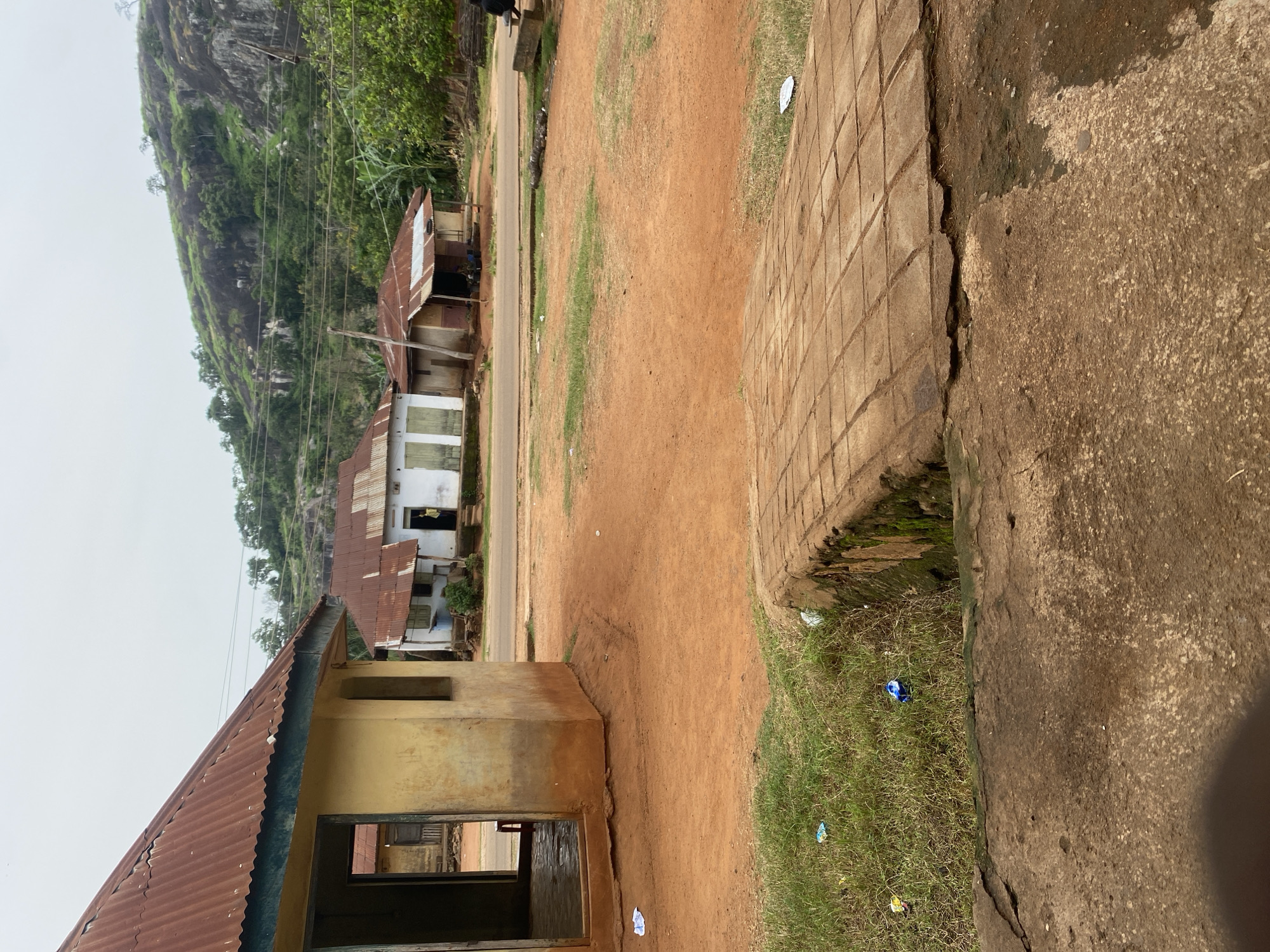 The image depicts a weathered, rural landscape with a prominent rocky hill covered in lush greenery in the background. In the mid-ground, several buildings with corrugated tin roofs are visible, some appearing to be in disrepair. A road runs between these structures and the foreground. The foreground is dominated by a dirt path and a rough, cracked concrete walkway bordering a patch of grass. The overall impression is one of a simple, perhaps impoverished, community situated in a natural, hilly environment. The scene is viewed from a slightly elevated perspective. The lighting suggests it is daytime, with overcast skies. There are no people visible in the frame, and the scattered litter on the grass and ground indicates a lack of maintenance.