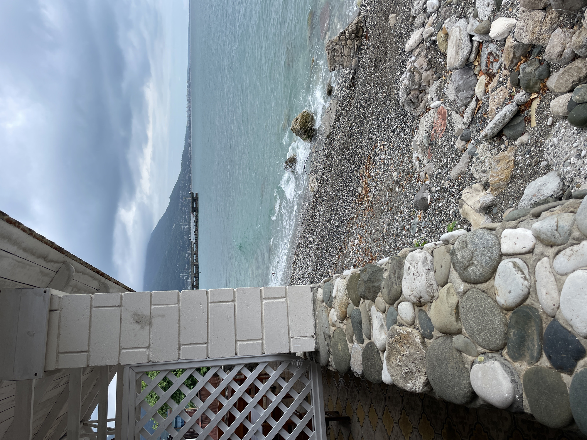 This is a photograph of a stony beach and the sea. In the foreground, a white lattice fence and a low wall made of large, rounded stones form a barrier. Beyond the wall, a pebble beach slopes down to the water. The sea is a muted blue-green, with small waves lapping at the shore and breaking around a few larger rocks. In the distance, a dark, tree-covered mountain rises steeply from the coastline. A pier or jetty extends into the water from the far shore. The sky is overcast with heavy, grey clouds, suggesting a cool or possibly rainy day. The overall mood is somewhat somber and tranquil. There are no people visible in the image. The lighting is diffused, indicating it is likely daytime, perhaps late afternoon or early morning, with the sun obscured by clouds. The location is identified as Abaata, Abkhazia.