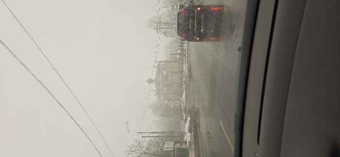 The image depicts an overcast, hazy winter or early spring day in Volosovo, Russia, viewed from the interior of a vehicle. The foreground shows the dashboard and the lower portion of the windshield. The asphalt road ahead is wet and reflective, with patches of dirty snow and slush along the shoulders.
In the midground, a dark-colored SUV, possibly a Hyundai Tucson, is prominently featured, facing away from the camera. Its red brake lights are illuminated, reflecting on the wet road surface. A lighter-colored vehicle is partially visible behind and to the right of the SUV. A green light, likely from a traffic signal, is visible to the right of the SUV.
The background is dominated by the Church of St. Nicholas, a light yellow or beige historical building with a distinctive green or grey dome and spire, characteristic of Russian Orthodox architecture. It is surrounded by a fence and bare trees. Utility poles with numerous overhead power lines crisscross the scene on the left, receding into the haze. Another, smaller building with a spire is partially visible to the far right, further obscured by the atmospheric conditions. No individuals are clearly visible.