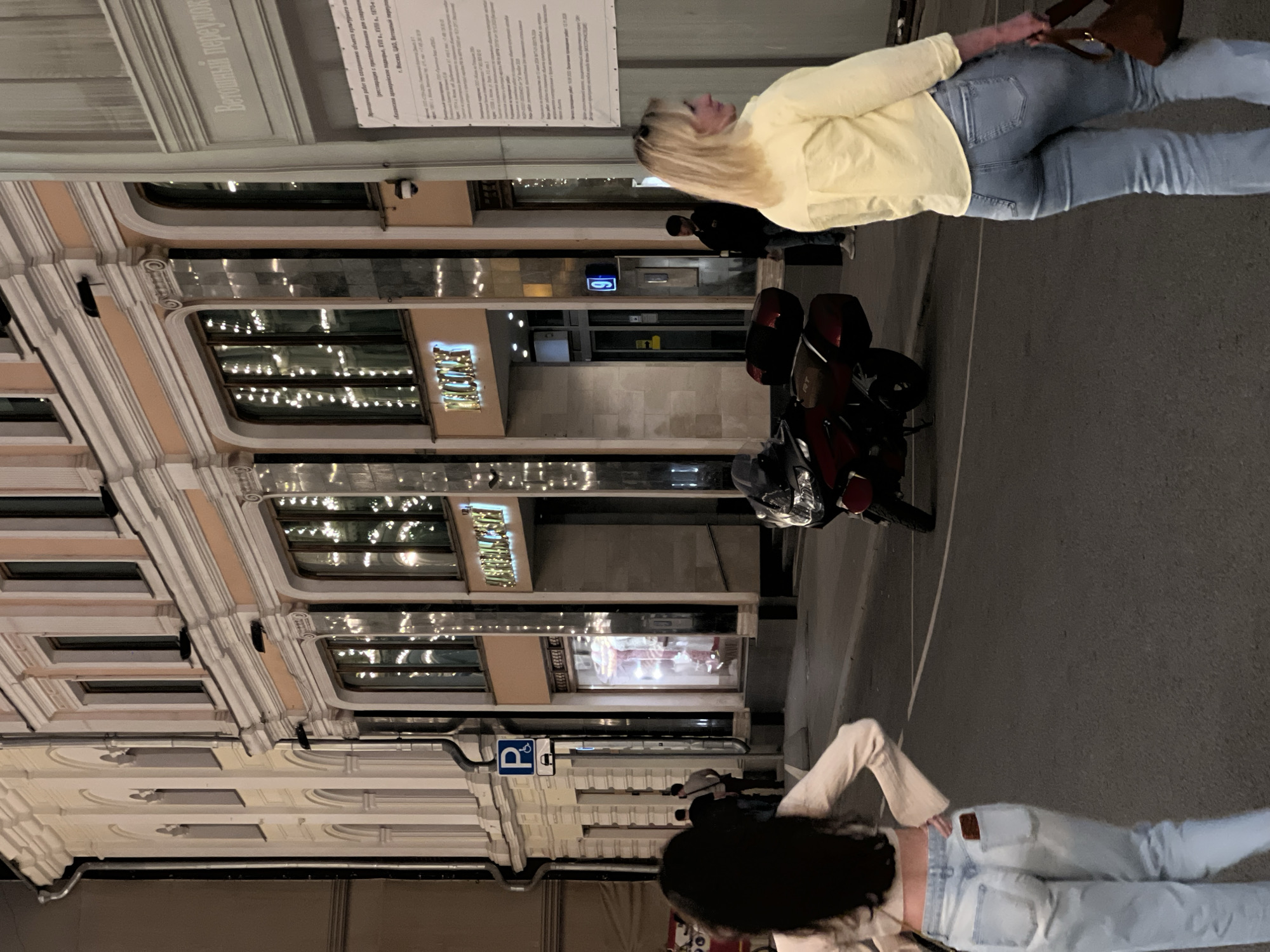 Here is a detailed description of the image:
The image captures a street scene in Moscow, Russia, specifically in the vicinity of Vetoshny Lane. The architecture features ornate building facades with decorative window frames, and festive lighting.
Two women are visible on the street. One woman, with blonde hair, is wearing a yellow jacket and jeans, and carrying a brown bag. The other woman, with long dark hair, is wearing a light-colored top and jeans.
A red motorcycle is parked on the street. Signs including a parking sign are visible.