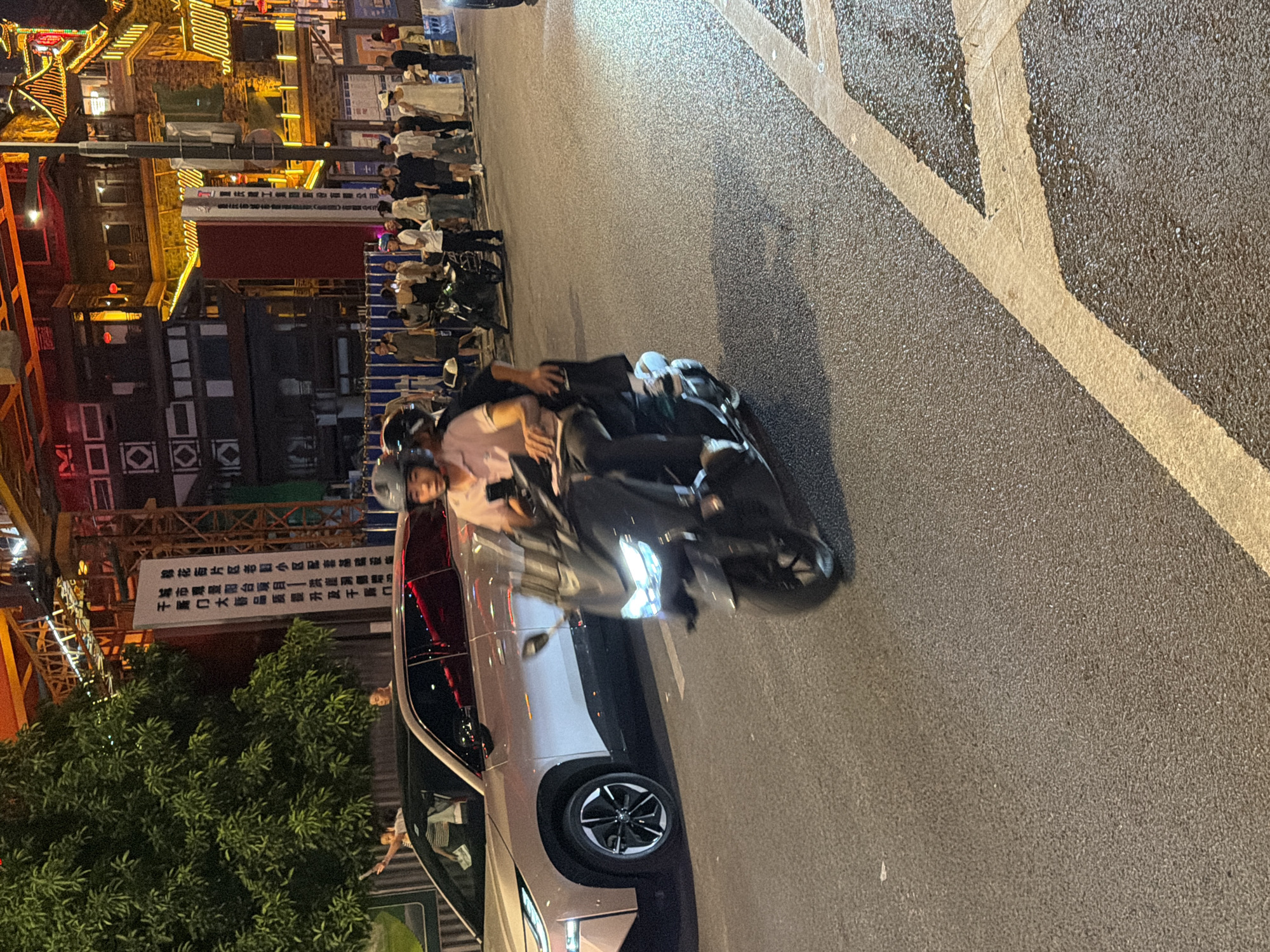 At night in Yuzhong District, China, on a paved road with yellow lane markings, two people ride on a dark-colored scooter. The driver, wearing a pink polo shirt and a helmet, holds a mobile phone. A passenger, wearing dark clothing, sits behind the driver. A light-colored SUV is parked to the left of the scooter. In the background, there is a large tree, construction cranes, and buildings illuminated with warm lights. A crowd of people, some dressed in white, are gathered on a sidewalk behind a temporary barrier. The scene appears to be a busy urban street at night.