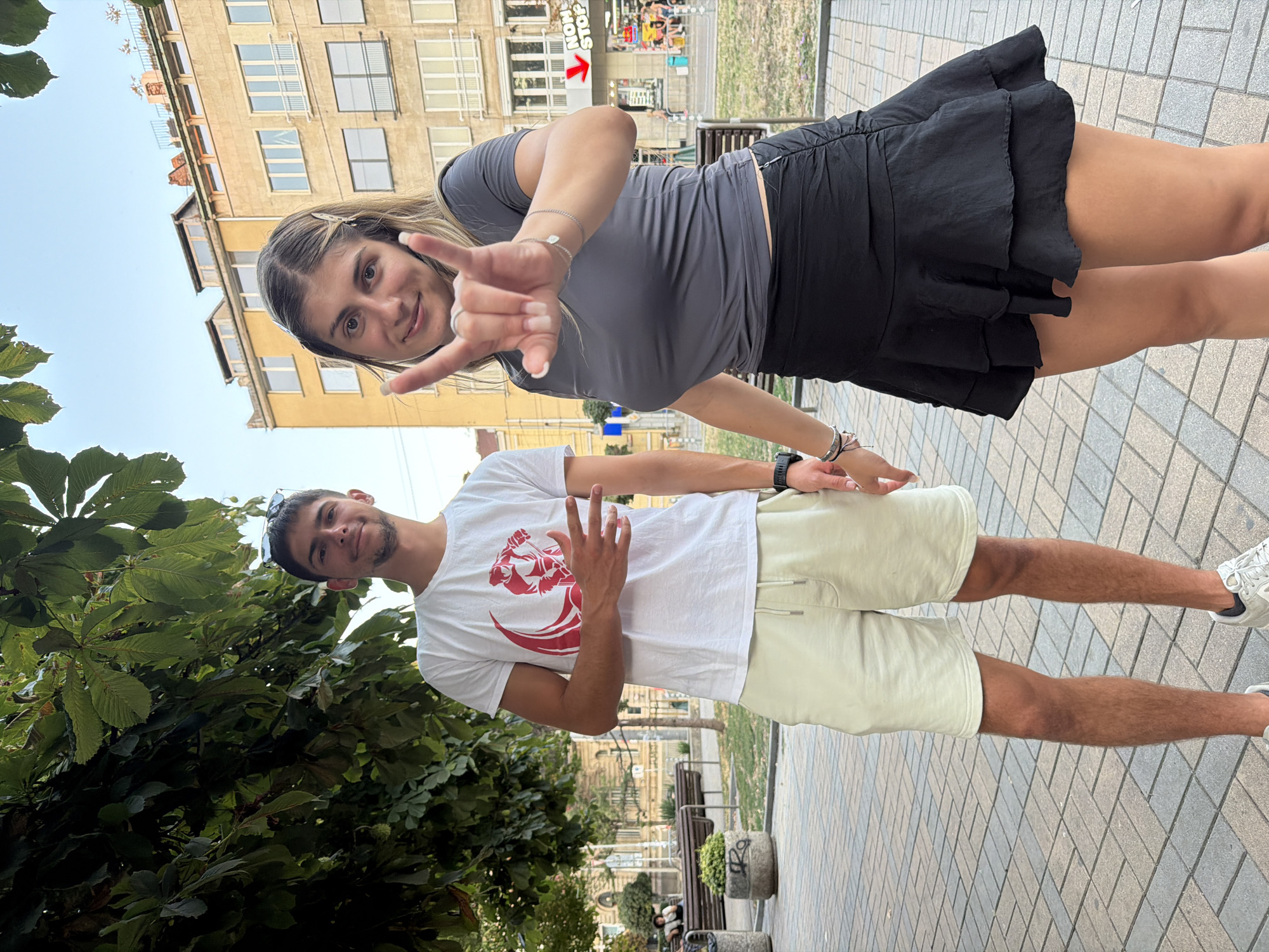 The image shows a young couple standing on a paved walkway in Sofia, Bulgaria. The man on the left is wearing a white t-shirt with a red graphic, light-colored shorts, and sneakers. He has short dark hair and is smiling at the camera, with his left hand held up in a gesture of peace or victory. The woman on the right is wearing a grey t-shirt and a black ruffled skirt. She has long blonde hair and is smiling at the camera, with her right hand raised in a rock-and-roll gesture. Behind them, there is a tree with lush green leaves, park benches, and a building with multiple windows and balconies. The overall scene appears to be a casual outdoor setting in an urban environment.