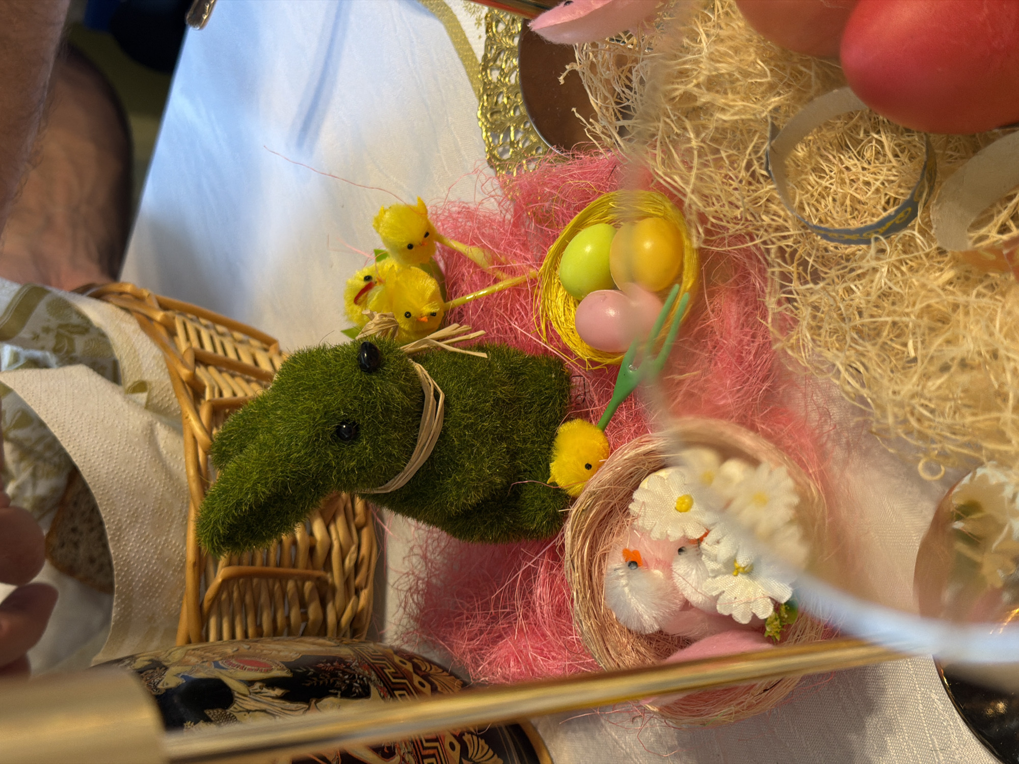 A close-up, high-angle shot shows an Easter-themed arrangement on a white tablecloth. A green moss-covered rabbit figure with black bead eyes and a straw tie is near a small wicker basket. Adjacent to the rabbit are clusters of yellow chicks and nests containing pastel eggs. One nest contains yellow, pink, and green eggs, while another nest houses miniature white birds decorated with small white flowers. The arrangement also includes a large nest made of straw-like material with pink and red Easter eggs. A decorative tray with colorful patterns is visible in the bottom left corner. A partial view of an arm is seen in the top left corner. The arrangement is positioned on a white tablecloth. There is no indication of Krasnoyarsk, Russia.