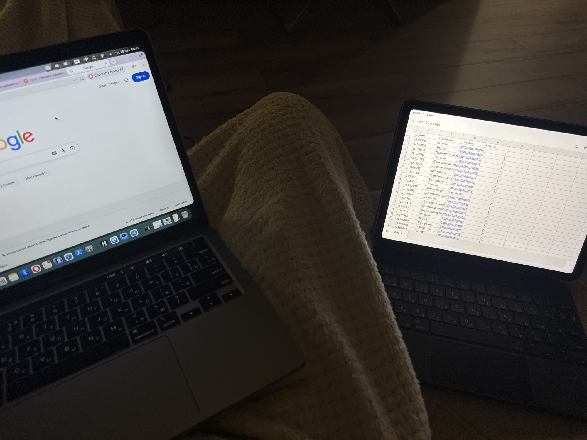This image shows a laptop and a tablet placed on top of what appears to be a blanket or padded surface. The laptop on the left displays the Google search engine homepage with Cyrillic script suggesting Russian localization. The tablet on the right shows a spreadsheet with labels like "Артикул" (Article), "Название" (Name), "Ссылки" (Links), and "Кол-во" (Quantity), indicating inventory or parts data, and a title indicating "John Deere". Both devices show the time "00:41" and the date "Чт, 28 мая" (Thursday, May 28th). Cyrillic keyboard layouts are visible on both devices. The surrounding environment appears dimly lit and residential. There is no indication that this scene is in Saint Petersburg.