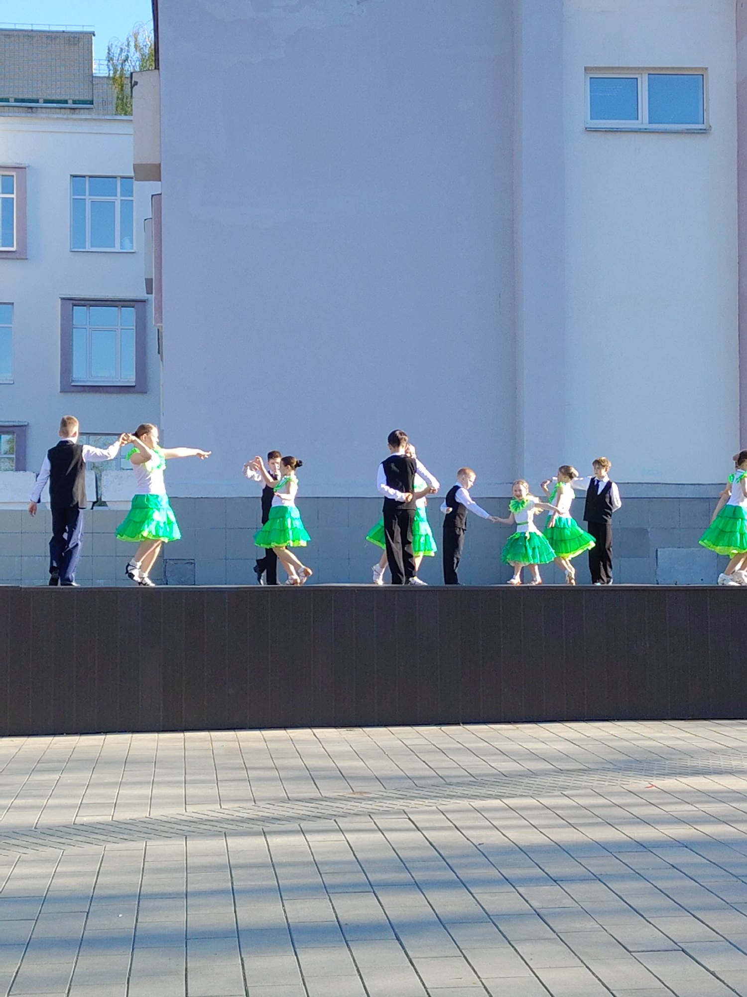 A group of young dancers are performing on a raised platform outdoors. The dancers, dressed in matching outfits consisting of white tops, green ruffled skirts, and vests, are arranged in pairs. They appear to be engaged in a coordinated dance routine. The platform is situated on a paved area consisting of rectangular stones. Behind the platform is a light-colored building with several windows. The sky is visible above the building. The location appears to be urban, potentially a town square or public space.