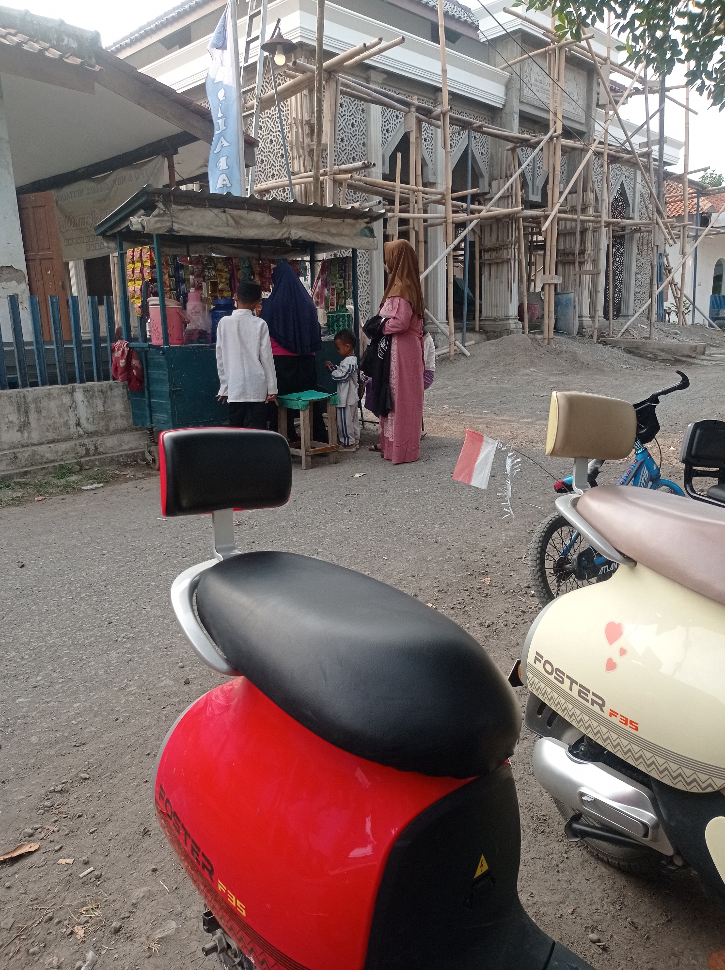 The image shows a street scene in Babakan, Indonesia, with a building under construction in the background, likely a mosque given the decorative elements. In the foreground, two motorcycles are parked, one red and one beige. A small street-side stall is visible on the left, selling various packaged goods. Several people are present, including a woman in a brown hijab, a child in a blue and white outfit, and a boy in a white shirt and black cap. A blue bicycle with a tan seat is also parked near the stall. A small Indonesian flag is attached to the bicycle. The ground is unpaved, with some debris visible. The overall atmosphere suggests a typical day in a village or small town setting.