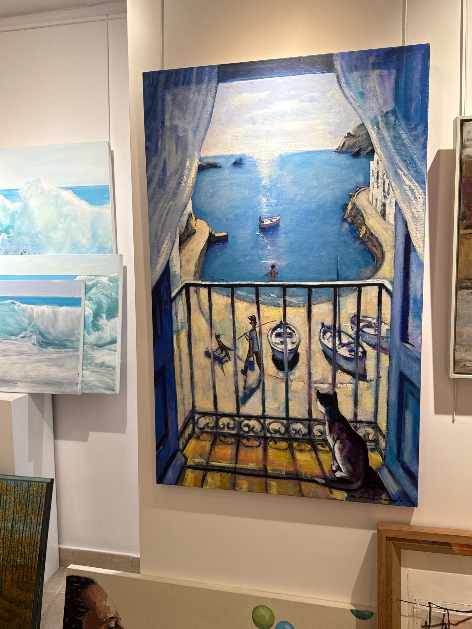 The image displays several paintings in what appears to be an art gallery setting. The most prominent painting is a large canvas depicting a view from a balcony overlooking a coastal town and the sea. The painting features a black and white cat sitting on the balcony, looking out at the scene. On the balcony, there are several boats and a figure walking with a bucket and fishing rod. The sea is depicted with boats and a person swimming, with buildings nestled into the cliffs.
To the left of this main painting, there are two smaller paintings leaning against each other, both depicting crashing waves and the ocean. In the lower left corner, a portion of another painting is visible, showing trees with autumnal foliage. Below these, a painting with a human face is partially visible. In the lower right corner, a framed artwork with abstract elements is present.
While the prompt specifies the city of Blanes, Spain, there are no explicit visual cues within the image itself (such as text, landmarks, or distinct architectural styles) that directly confirm this location. The scene depicted in the main painting, with its Mediterranean-style architecture and coastal setting, is consistent with many seaside towns in Spain, including potentially Blanes, but this cannot be definitively determined from the visual information alone.
