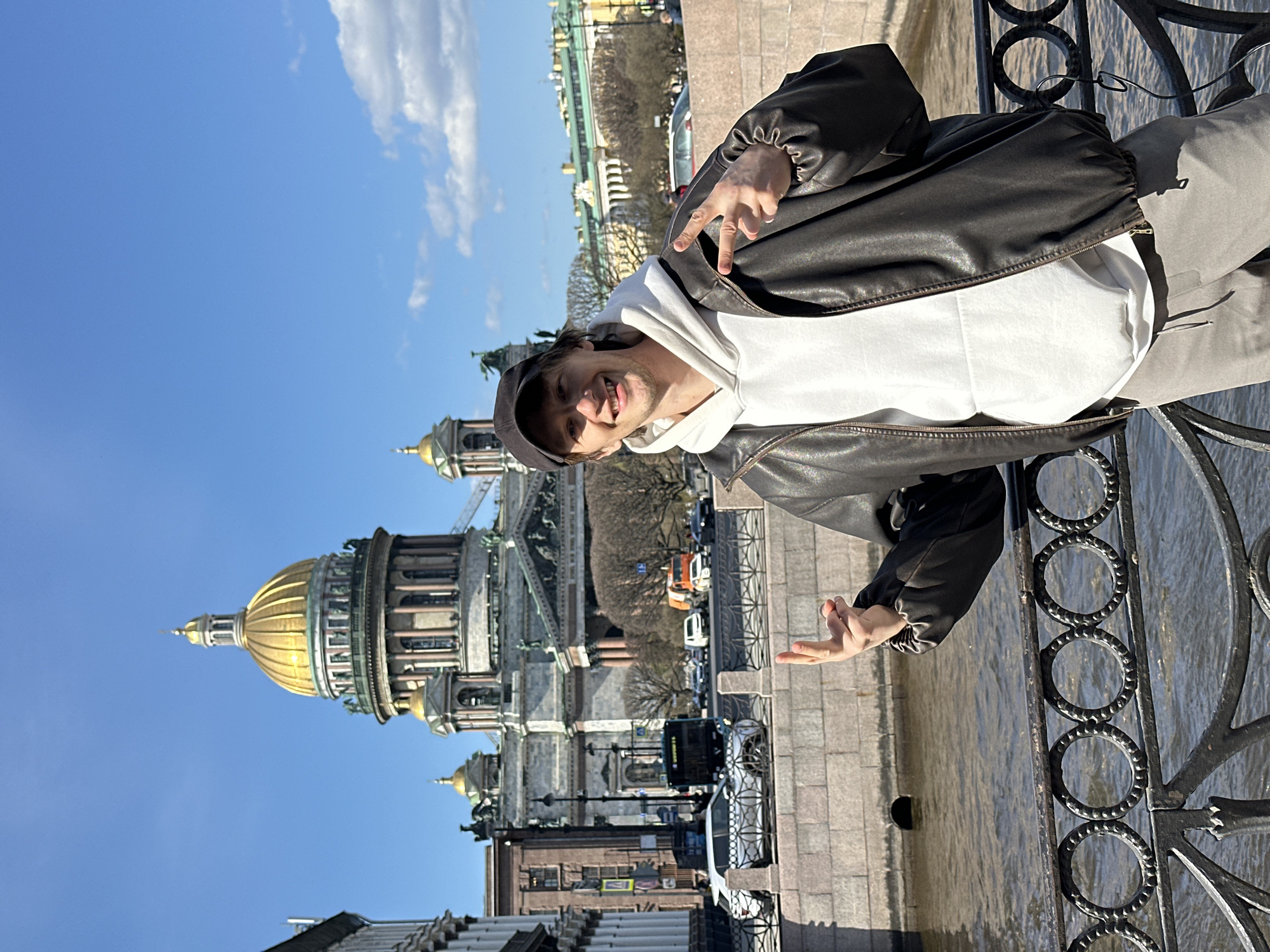 A young man is featured in a sunlit outdoor shot, posing with a large architectural landmark in the background. He is positioned on the right side of the frame, smiling broadly and looking towards the camera. He wears a black leather jacket over a white hooded sweatshirt, light grey pants, and a dark baseball cap. His right hand is raised, forming a 'V' or peace sign, while his left hand is held lower with an open palm.
Behind him, on the left, is the iconic St. Isaac's Cathedral in Saint Petersburg, Russia, easily recognizable by its massive golden dome and neoclassical columns. The building's intricate details are visible, including statuary and multiple levels of architecture. The sky above is a clear, bright blue with a few scattered white clouds, indicating fine weather.
To the man's right, a decorative black wrought iron railing separates the foreground from a body of water, likely one of Saint Petersburg's canals or rivers, which appears calm. Across the water, further into the background, are other urban buildings, some bare trees, and vehicles parked along a street. The bright natural light suggests it is daytime, possibly mid-morning or afternoon. No discernible text is visible on buildings or signs.