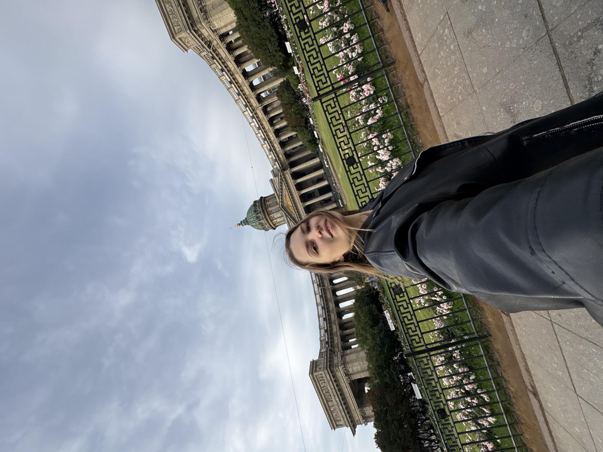 A young woman is taking a selfie with the Kazan Cathedral in Saint Petersburg, Russia, in the background. She is wearing a black leather jacket. The cathedral is a large, neoclassical building with a semicircular colonnade, a dome, and intricate architectural details. In front of the cathedral, there is a park with manicured lawns, rose bushes in bloom, and black metal fences. The sky is overcast.