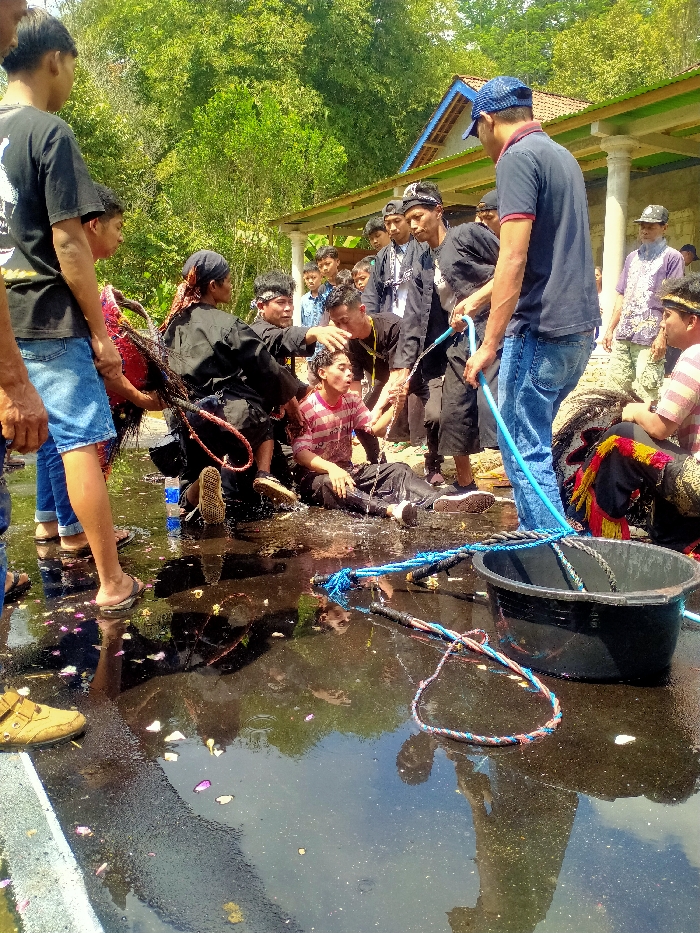 In Ngargoyoso, Indonesia, a public outdoor scene captures a ritualistic or performance event on a sunlit paved area, largely covered by a reflective puddle containing scattered pink and white petals. In the center, a young male in a red and white striped shirt and dark pants is seated or kneeling in the shallow water, his mouth open. He is being doused with water from a bright blue hose held by a man in a blue cap, blue t-shirt, and jeans standing to the right. To the left of the central figure, a person in dark attire and a headscarf kneels in the water, holding a bundle of dark, tangled ropes or strands. Another individual in dark clothing stands behind the seated male, placing a hand on his head.
Numerous other individuals, predominantly males, observe the activity from various positions around the central action. A large black rectangular bucket is situated in the foreground to the right, partially submerged in the water, near several blue and multicolored braided ropes. In the background, lush green foliage and a white building with a blue-tiled roof and pillars are visible. The ground is wet and reflective, indicating active water use, with reflections of the participants visible in the puddle.