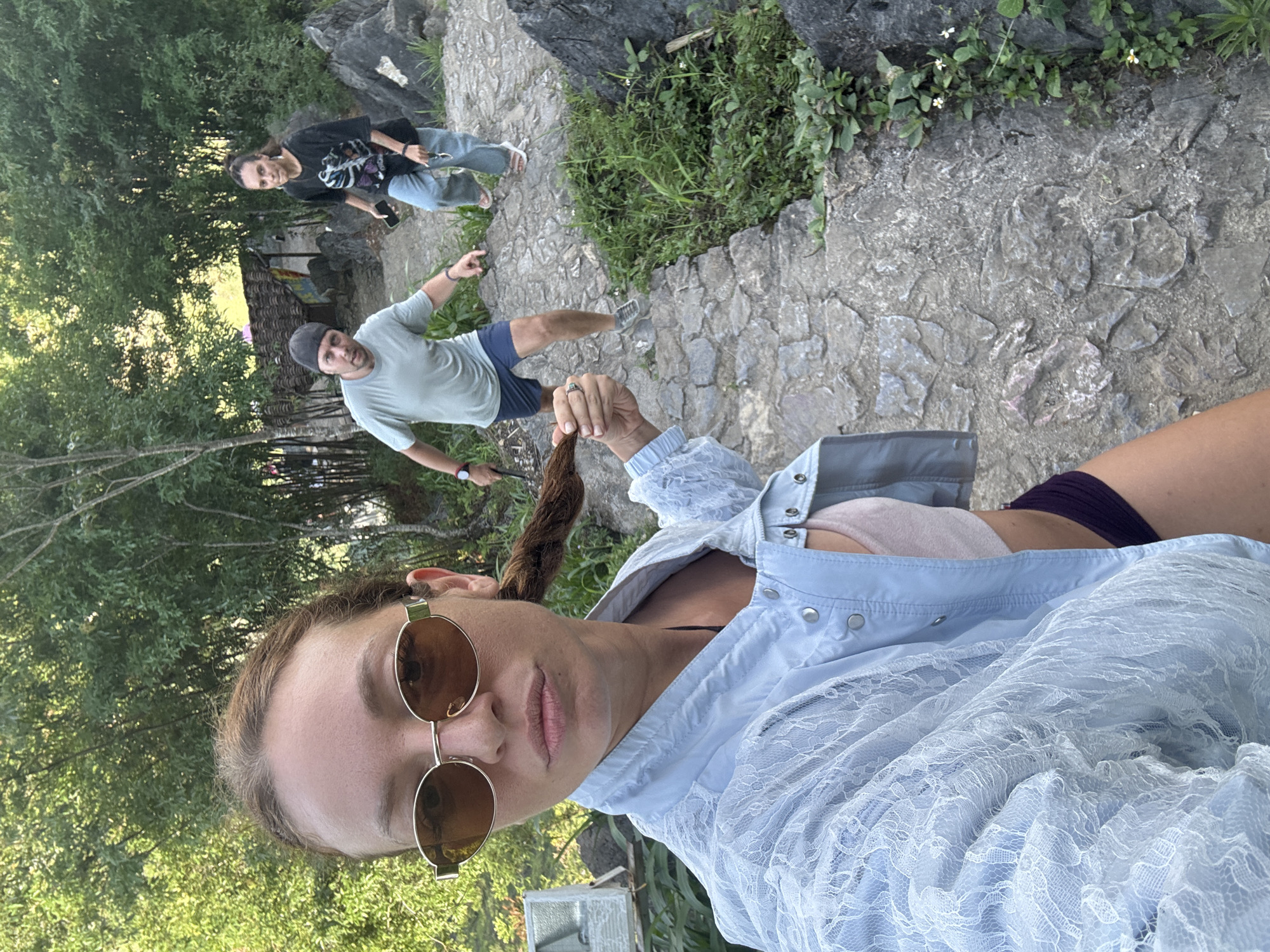 The image, taken at a high angle, shows a selfie of a person wearing round sunglasses and a light blue lace jacket. The person is holding a lock of hair. In the background, there are two other individuals on a stone path: one wearing a black t-shirt and jeans, and another in a light green t-shirt and blue shorts, pointing. The surrounding environment includes trees, stone structures, and vegetation. The location is in Đồng Văn Commune, Vietnam.