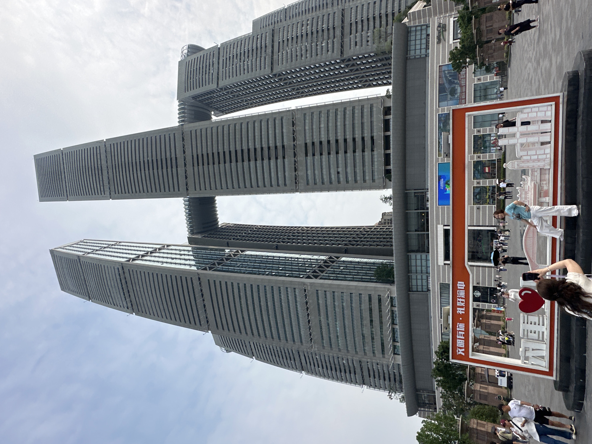 The image shows the Raffles City Chongqing complex, specifically a view of the skybridge linking the skyscrapers. Several people are present, some taking photographs near a large decorative sign with Chinese characters. The sky is cloudy. The location is in the Nan'an District, Chongqing, China.