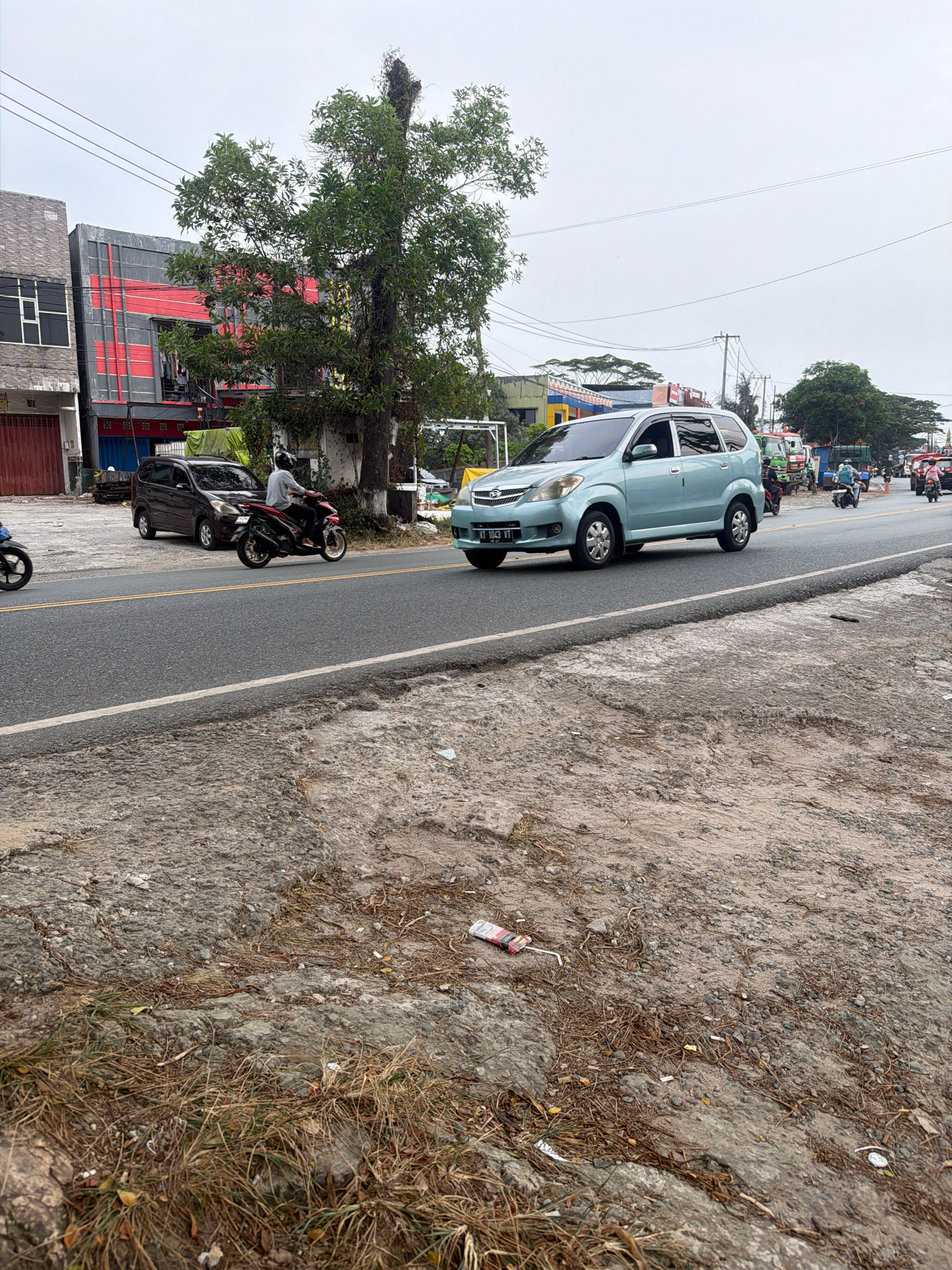 The image shows a street scene in Balikpapan, Indonesia. A light blue MPV is driving on the right side of the road, in the lane closest to the camera. A motorcycle with a rider wearing a white shirt and helmet is also on the road, to the left of the MPV. A dark colored SUV is parked on the side of the road, behind the motorcycle. In the background, there are several buildings, including a multi-story building with red and grey accents, and other structures that appear to be shops or businesses. Trees and utility poles are also visible. The ground in the foreground is a mix of dirt, gravel, and dry grass, with some litter scattered around. The sky is overcast. The road is paved and appears to be in moderate condition with some patches and cracks. The scene depicts typical urban traffic and streetscape in Indonesia.